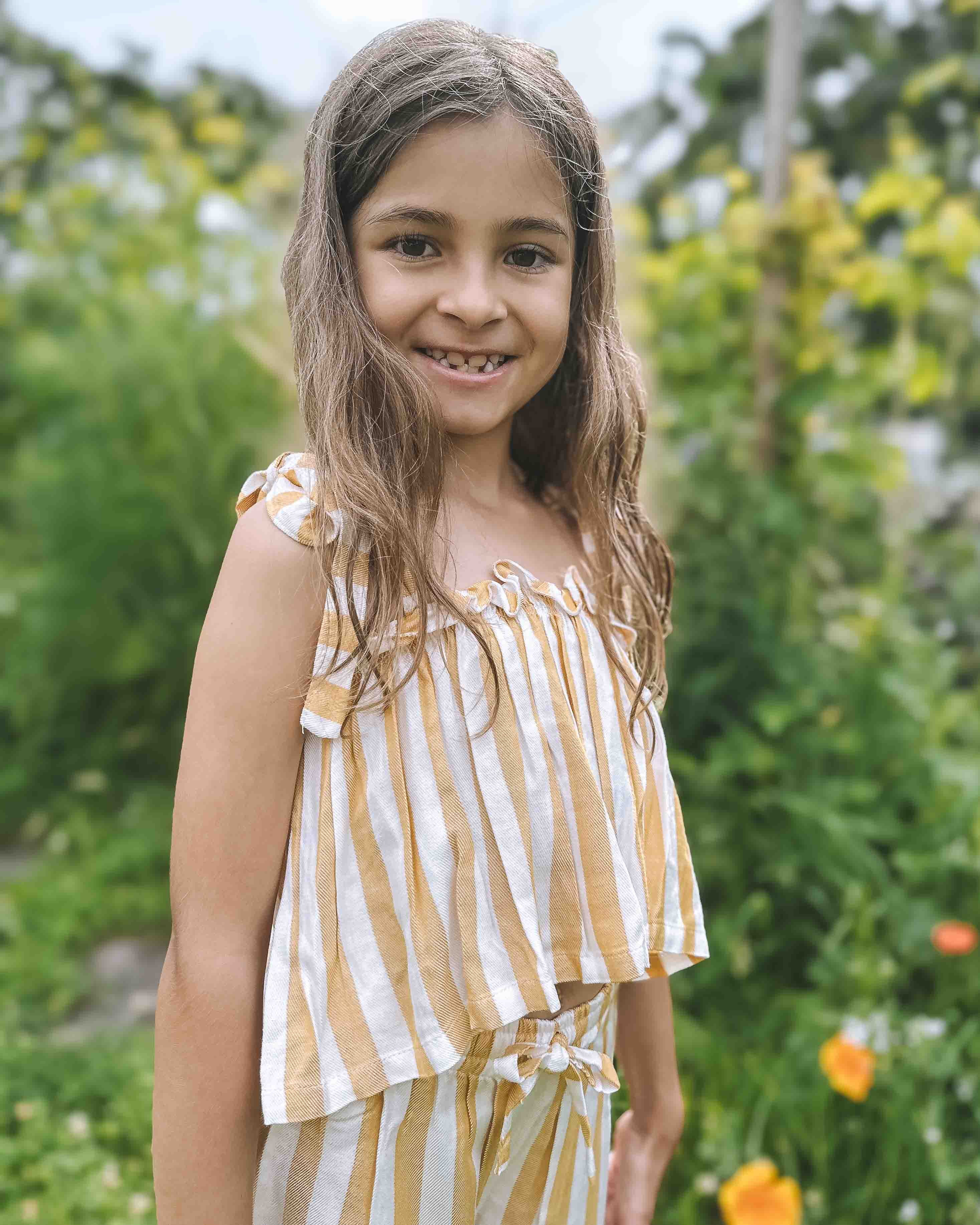 Lächelndes junges Mädchen mit langen braunen Haaren trägt das Main Design Boho Hawaii Set, ein gelb-weiß gestreiftes Outfit, während sie draußen in einem üppigen grünen Garten mit orangefarbenen Blumen und verschwommenem Laub im Hintergrund steht.