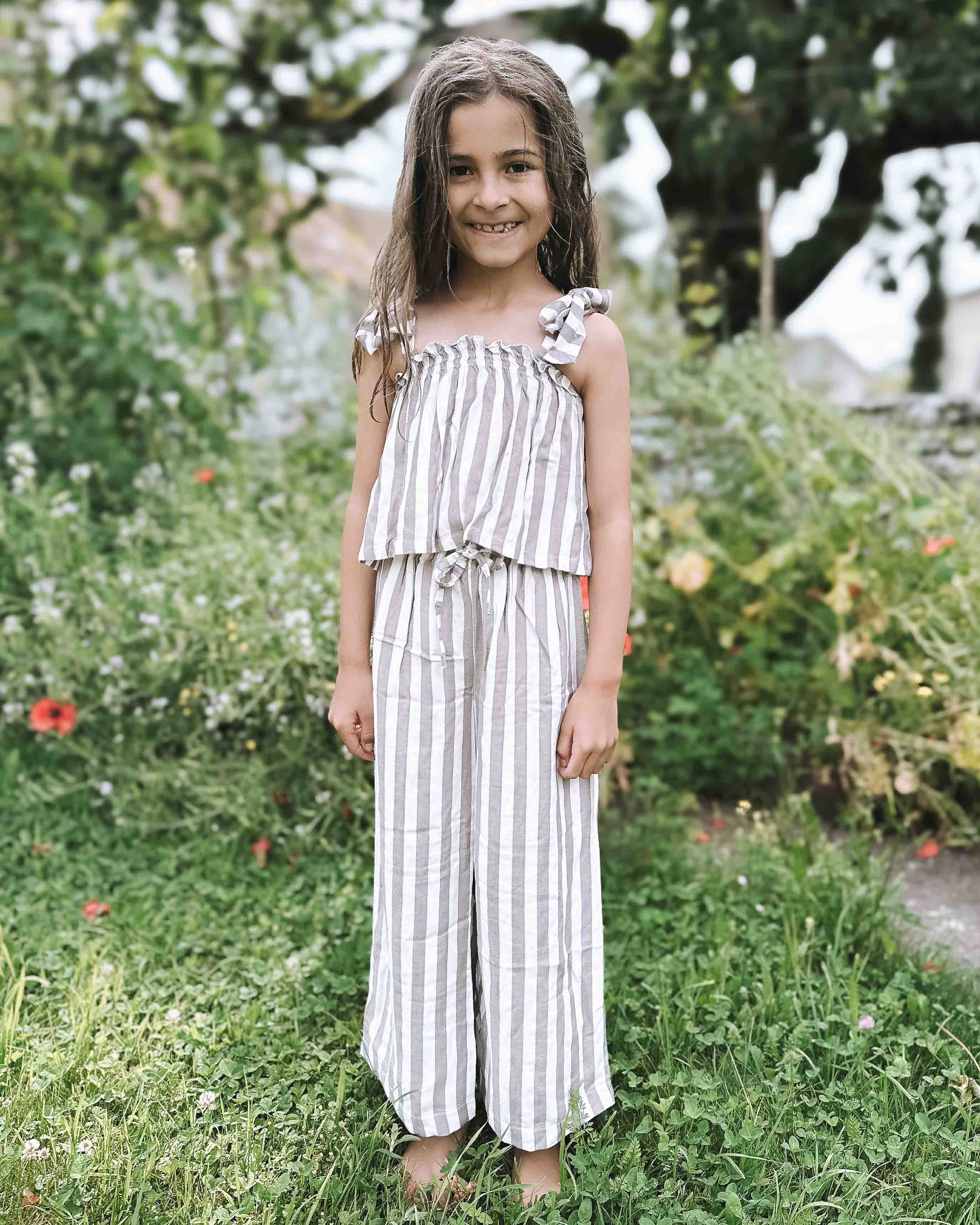 Ein junges Mädchen mit langen braunen Haaren steht barfuß lächelnd im Gras. Sie trägt das Boho Hawaii Set von Main Design, einen Zweiteiler im Boho-Stil mit Rüschendetails aus hochwertiger Viskose. Hinter ihr erstreckt sich ein üppiger Garten voller Pflanzen und Blumen.