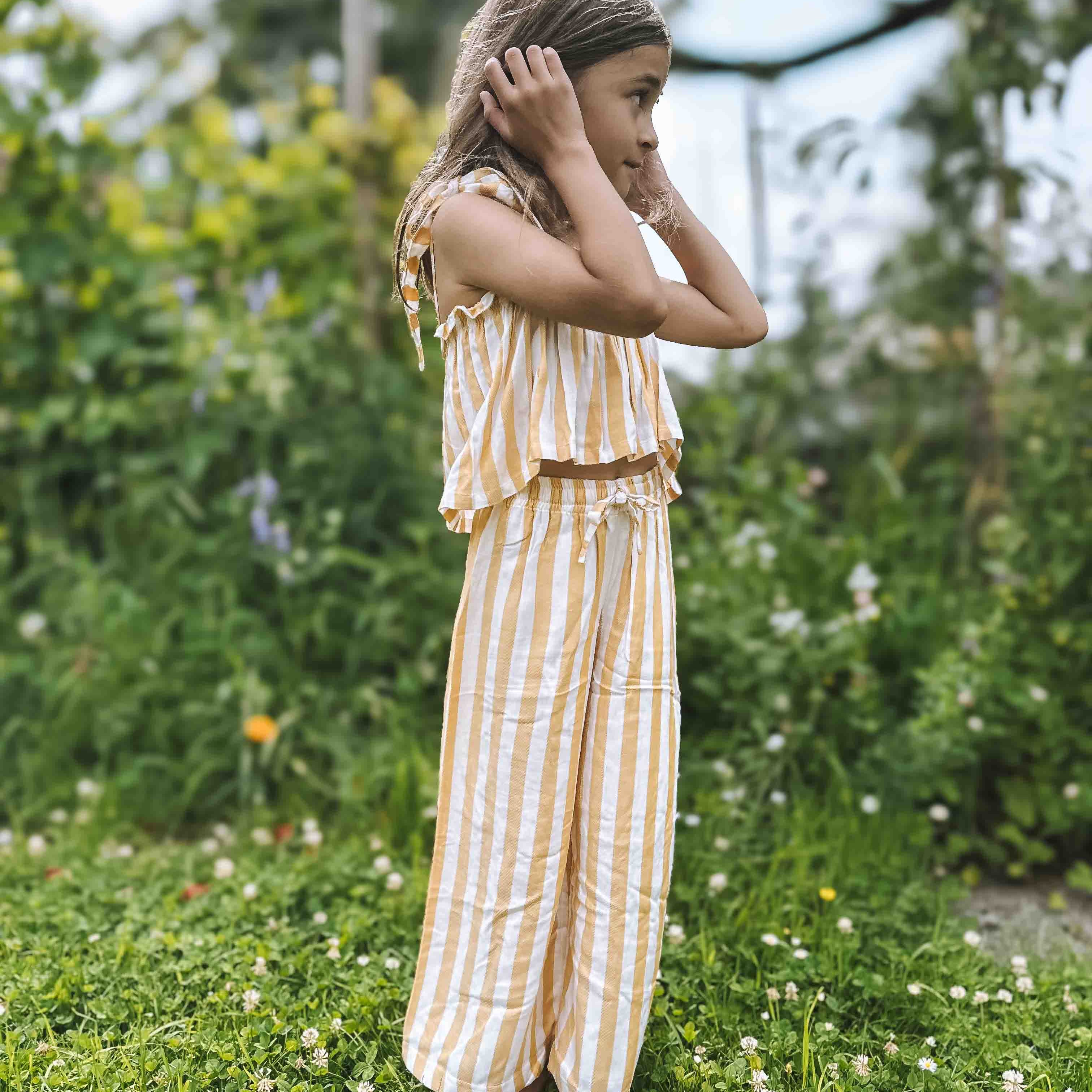 Ein junges Mädchen mit hellbraunem Haar steht barfuß im Gras eines Gartens und trägt das Boho Hawaii Set von Main Design. Sie hält ihre Hände an den Ohren und blickt zur Seite vor einem Hintergrund aus grünen, blattreichen Pflanzen.