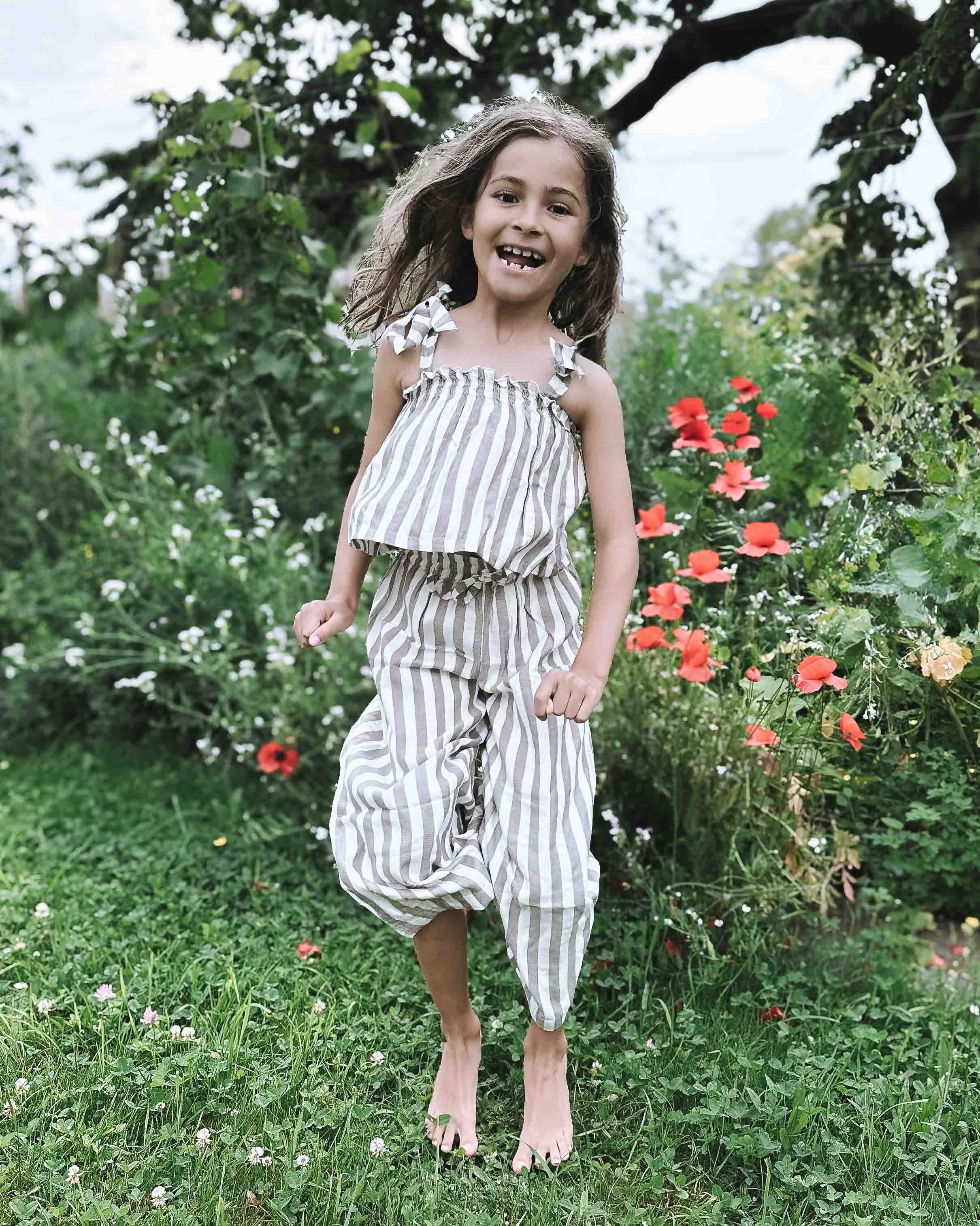 Ein lächelndes junges Mädchen mit langen Haaren steht barfuß auf dem Gras in einem Garten und trägt das Boho Hawaii Set von Main Design, mit grünen Pflanzen und roten Blumen im Hintergrund.