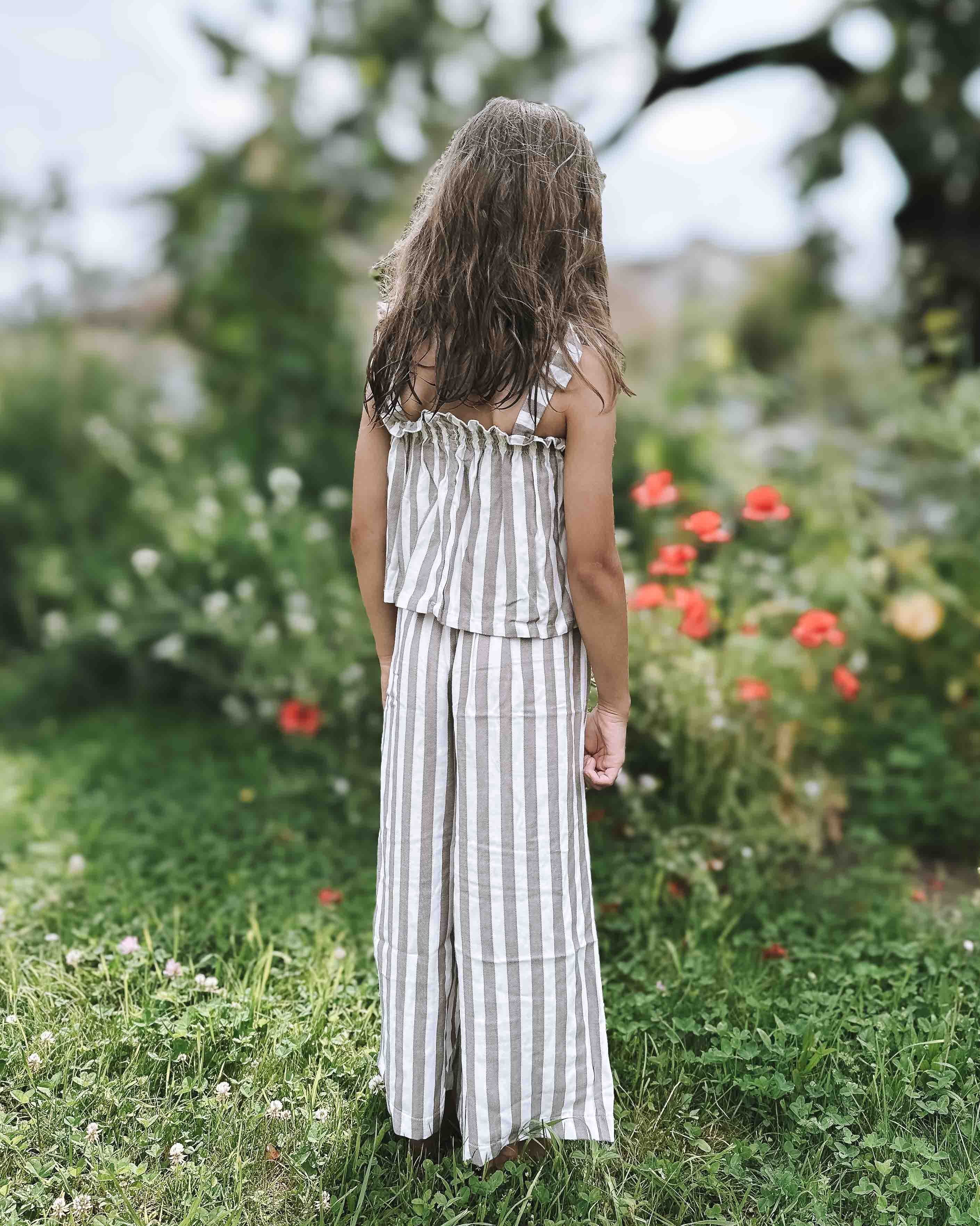 Ein junges Mädchen mit langen braunen Haaren, gekleidet im Boho Hawaii Set von Main Design, steht mit dem Rücken zur Kamera in einem Garten voller grünem Gras und roten Blumen.