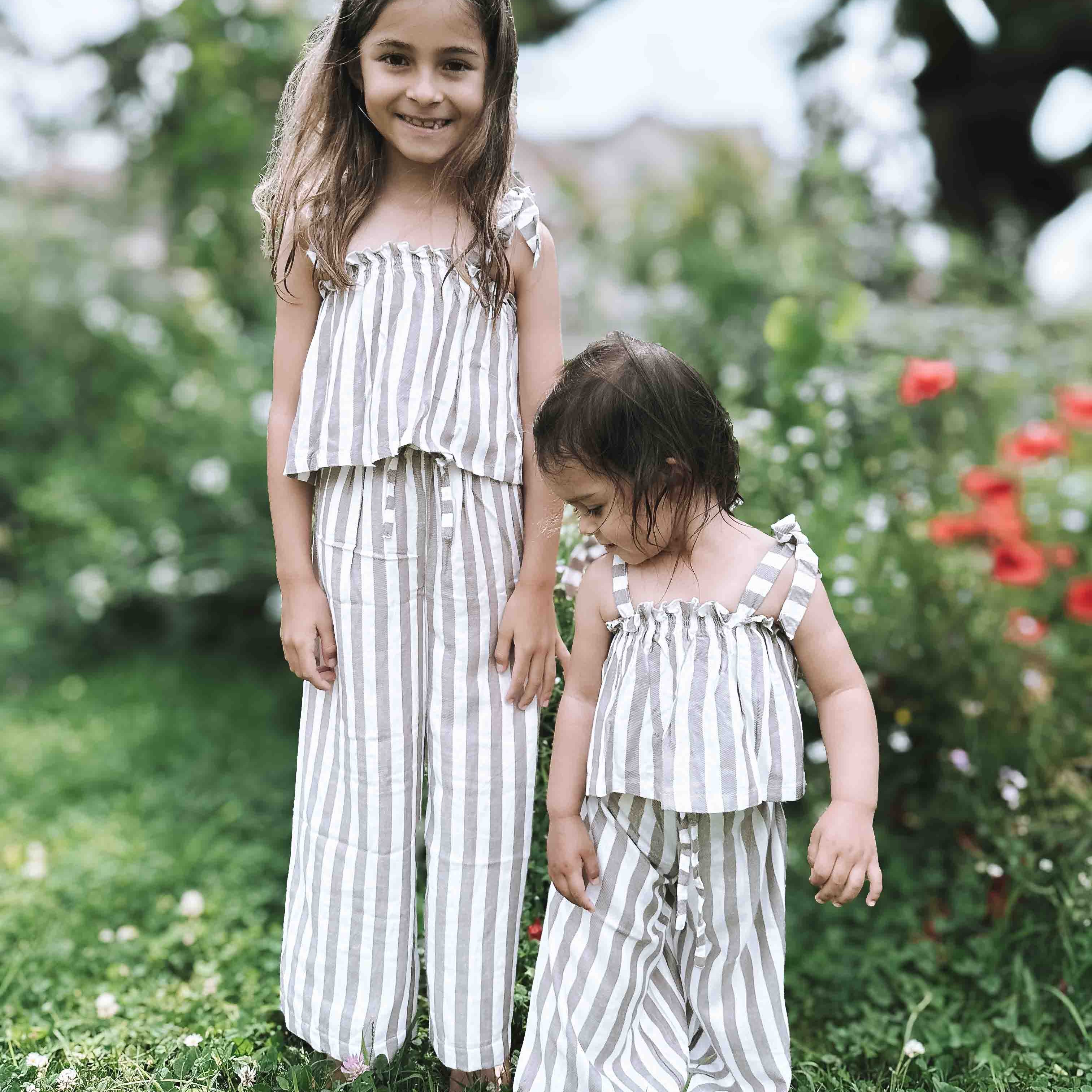 Zwei kleine Kinder stehen draußen auf der Wiese. Beide tragen passende hell gestreifte Outfits aus dem Main Design Boho Hawaii Set. Das ältere Mädchen lächelt in die Kamera, während leuchtende Blumen und Grün sie in ihrem stilvollen Set umgeben.