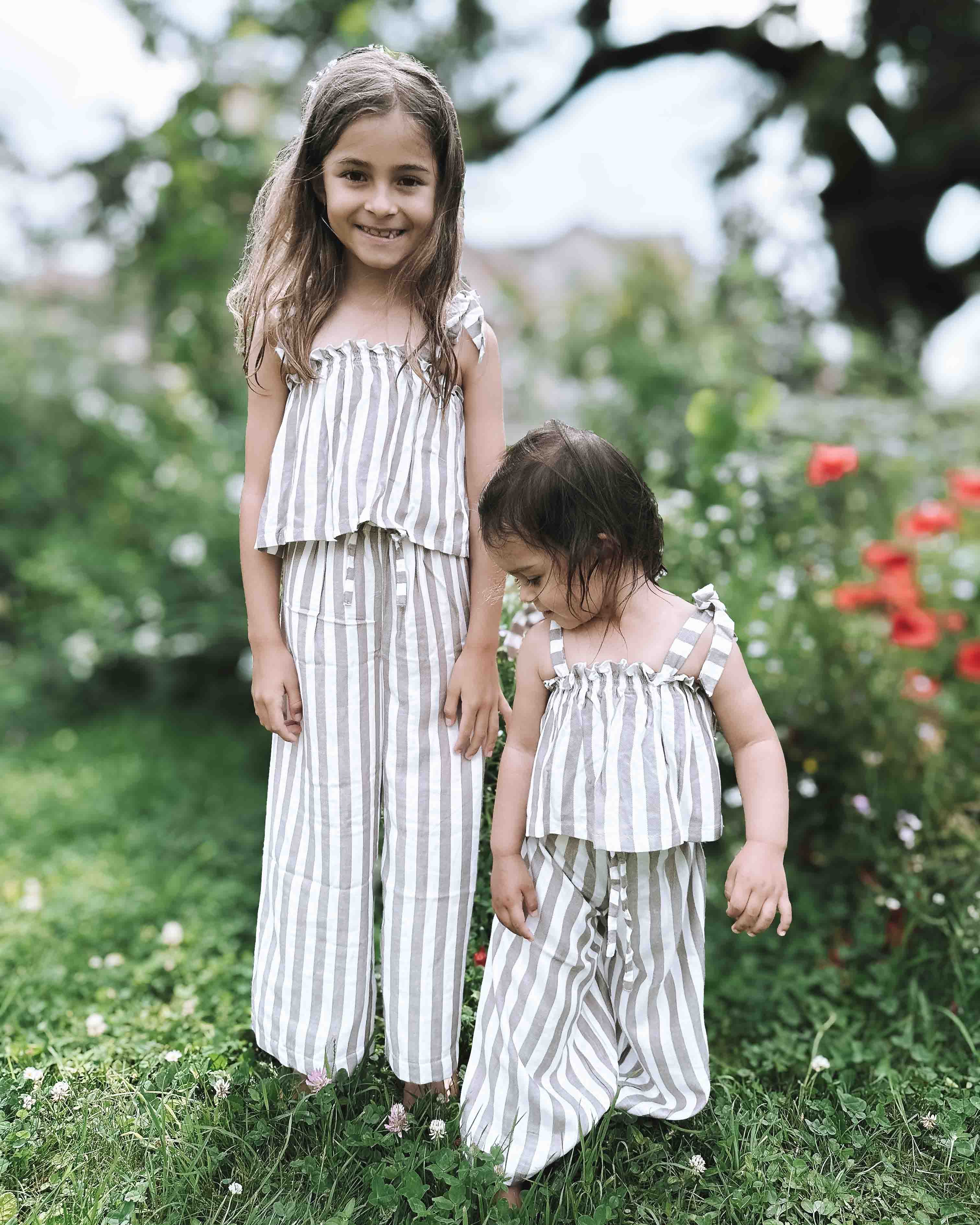 Zwei kleine Kinder stehen draußen auf der Wiese. Beide tragen passende hell gestreifte Outfits aus dem Main Design Boho Hawaii Set. Das ältere Mädchen lächelt in die Kamera, während leuchtende Blumen und Grün sie in ihrem stilvollen Set umgeben.