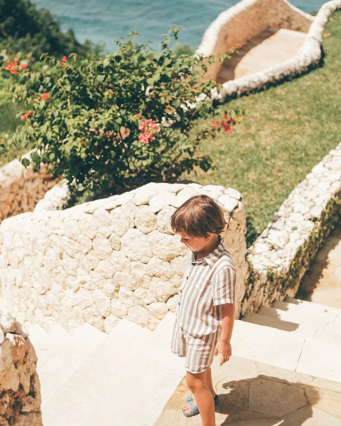 Ein kleines Kind in einem Boho-Shirt-Set von Main Design geht Steintreppen hinunter, die von weißen Wänden und grünen Pflanzen gesäumt sind, im Hintergrund ist das Meer zu sehen.