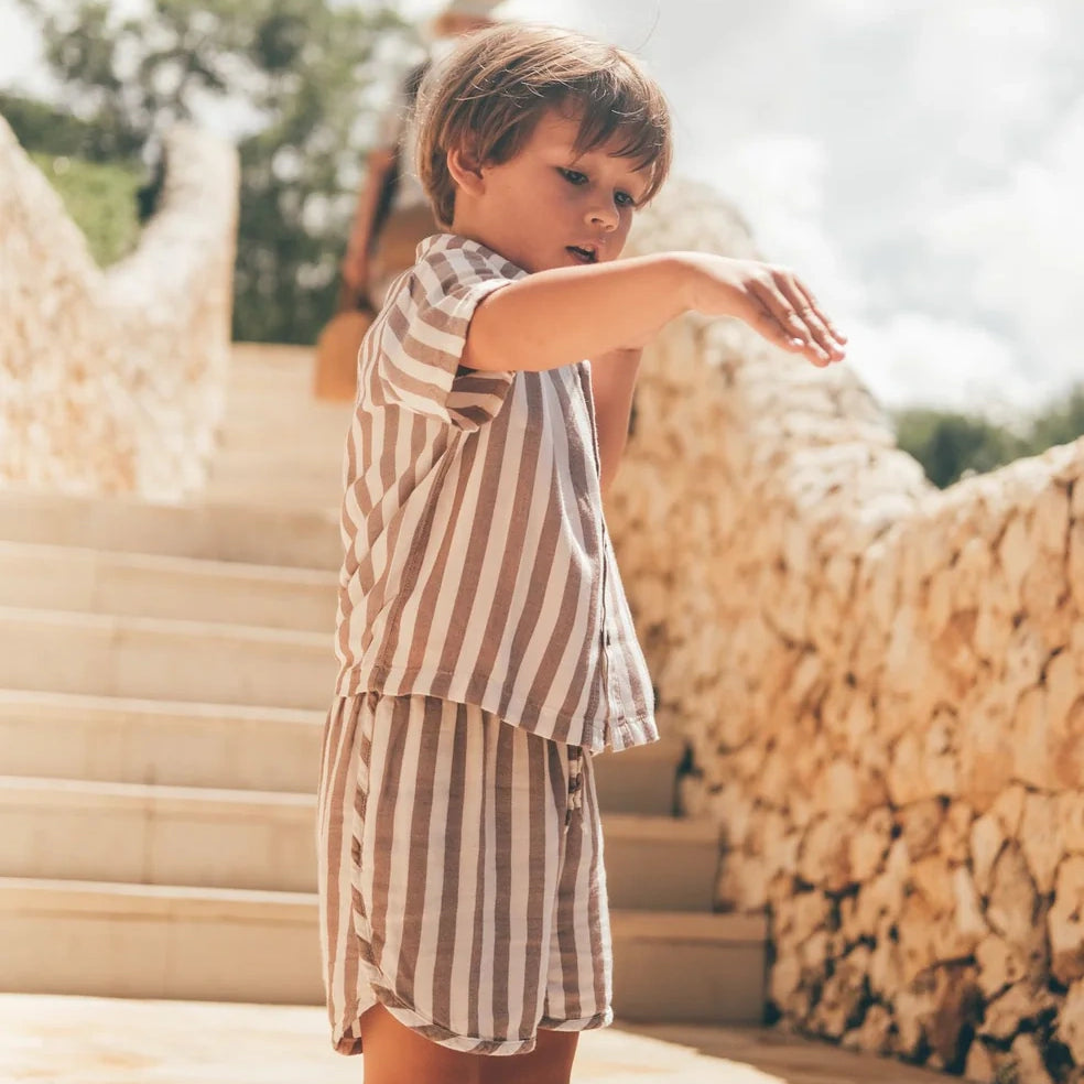 Ein kleines Kind in einem Boho-Shirt-Set von Main Design, ideal für Kinder-Sommerkleidung, steht mit ausgestrecktem Arm auf sonnigen Steinstufen. Dahinter sind Steinmauern und Grünflächen unter einem teilweise bewölkten Himmel zu sehen.