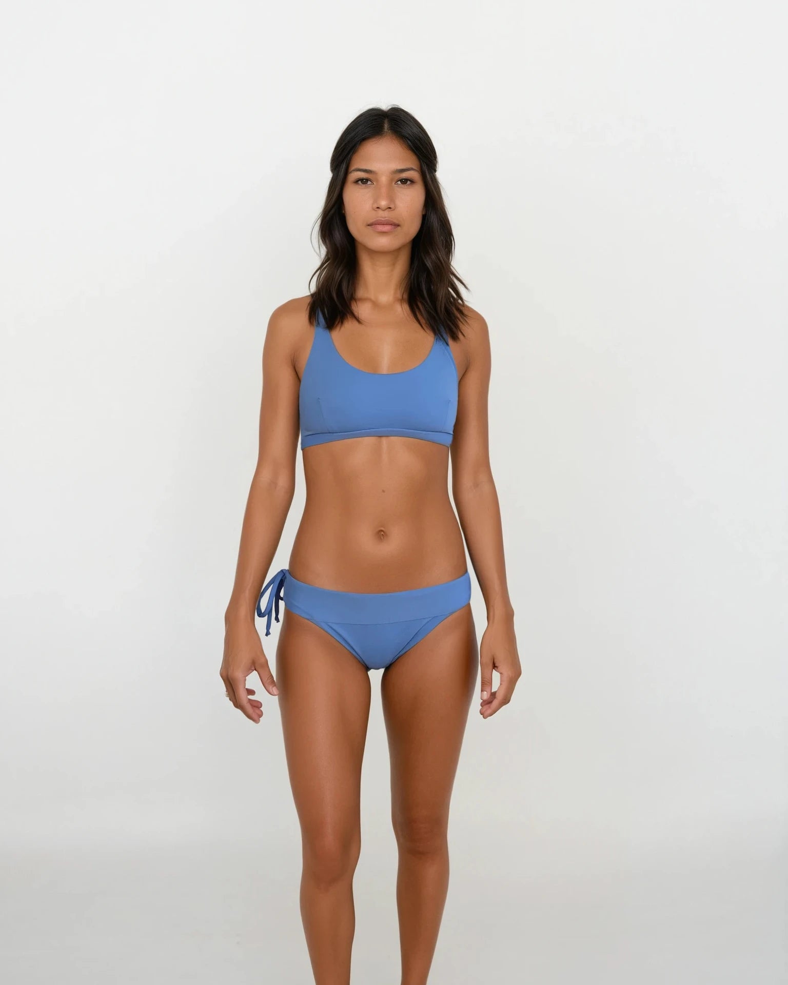 Eine Frau mit glattem, dunklem Haar und neutralem Gesichtsausdruck steht vor einem weißen Hintergrund. Sie trägt das Main Design Charm Surf Bikini Oberteil und eine blaue Hose mit seitlichen Bindebändern. Das Set bietet LSF 50+ Sonnenschutz – ideal für Wassersport.