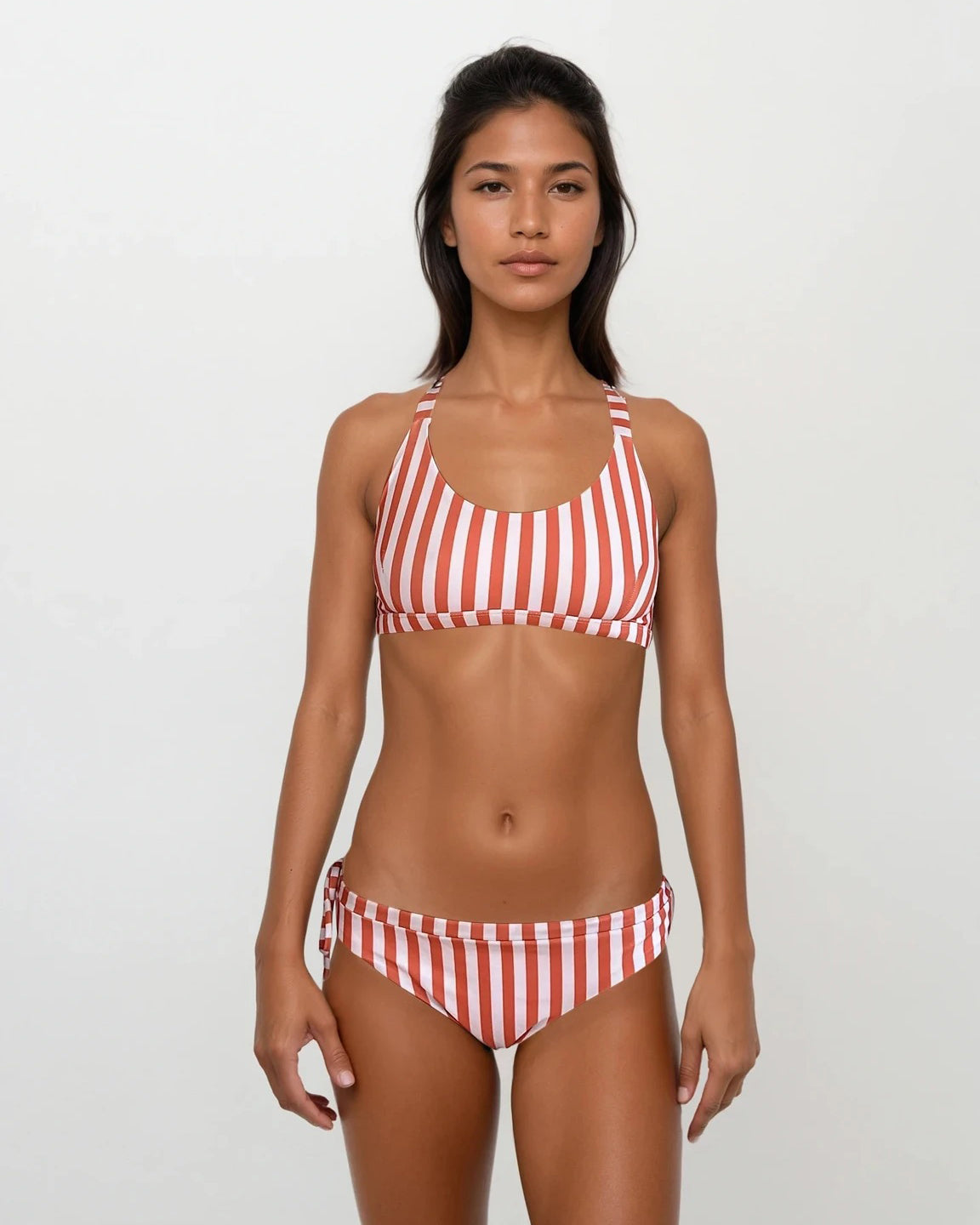 Eine Frau steht vor einem schlichten hellen Hintergrund. Sie trägt einen rot-weißen, vertikal gestreiften Bikini mit sportlichem Oberteil und einer passenden Main Design Blush Surf Bikinihose. Sie blickt mit neutralem Gesichtsausdruck direkt in die Kamera.
