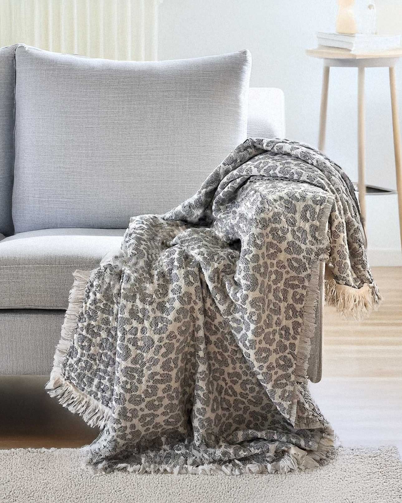 Die Decke Savanna von Main Design mit grauem Leopardenmuster und fransigen Rändern wird in einem modernen Wohnzimmer neben einem kleinen runden Holztisch über die Armlehnen und die Sitzfläche eines hellgrauen Polstersessels drapiert.