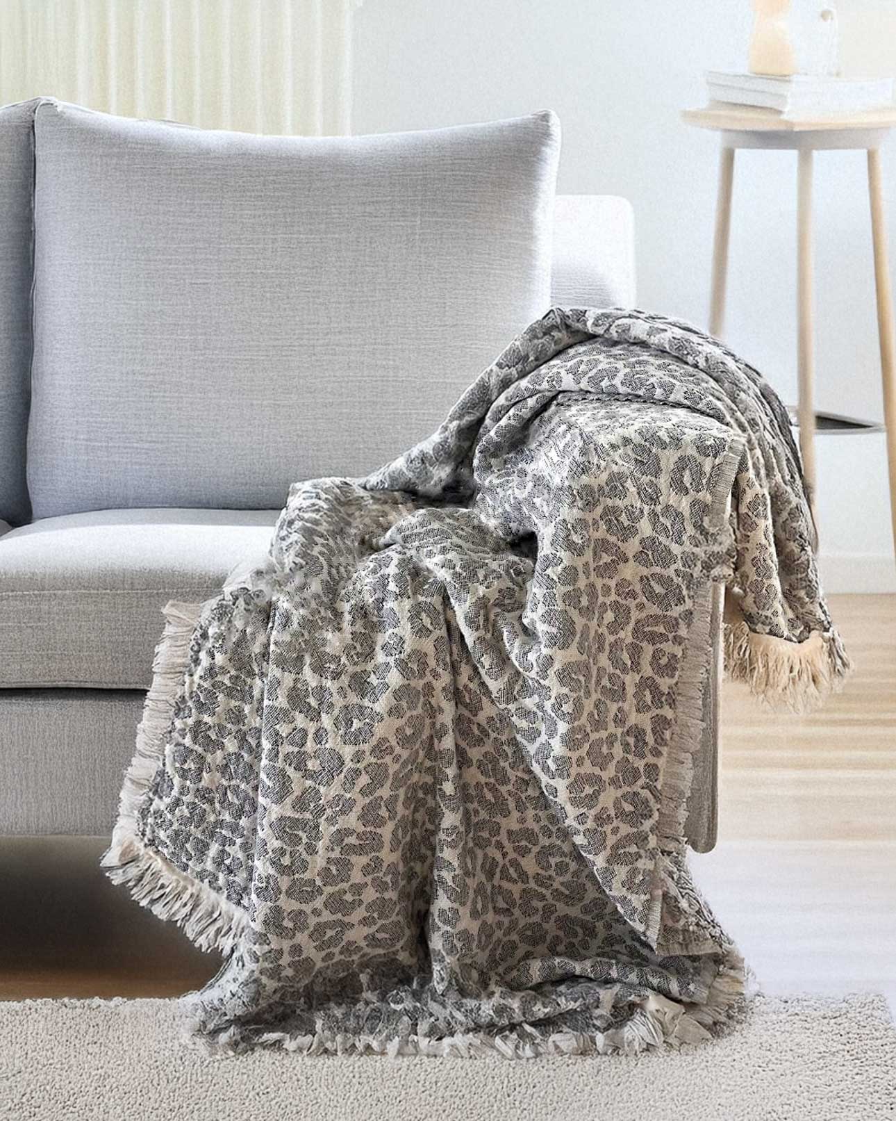 Die Decke Savanna von Main Design mit grauem Leopardenmuster und fransigen Rändern wird in einem modernen Wohnzimmer neben einem kleinen runden Holztisch über die Armlehnen und die Sitzfläche eines hellgrauen Polstersessels drapiert.