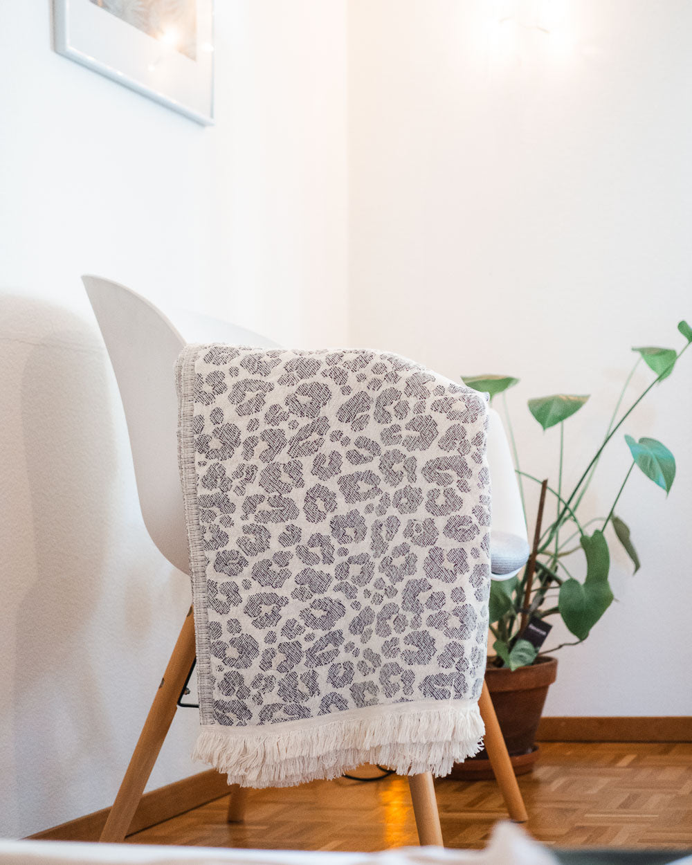 Auf einem weißen Stuhl mit Holzbeinen liegt die Main Design Decke Savanna, eine grau-weiß gemusterte Decke, die über der Rückenlehne drapiert ist. Im Hintergrund steht eine Topfpflanze auf einem Holzboden in einem hellen, minimalistischen Raum.