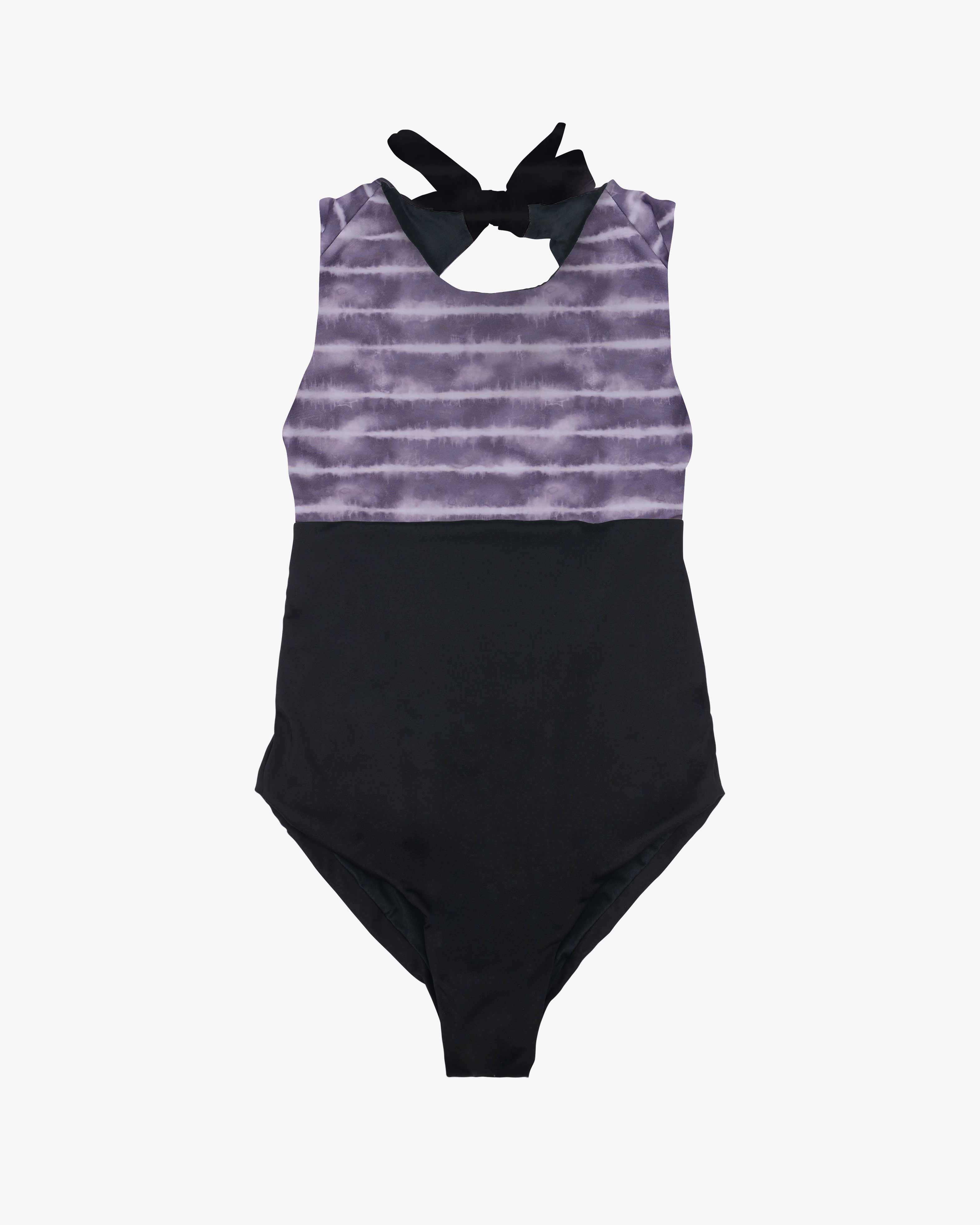 Der Divine Surf Badeanzug von Main Design ist ärmellos und besticht durch ein auffälliges lila Batik-Oberteil und ein schwarzes Unterteil. Er hat einen Neckholder mit schwarzer Binderückseite und ist aus umweltfreundlichem Stoff gefertigt, was ihn zu einer umweltfreundlichen Wahl macht.