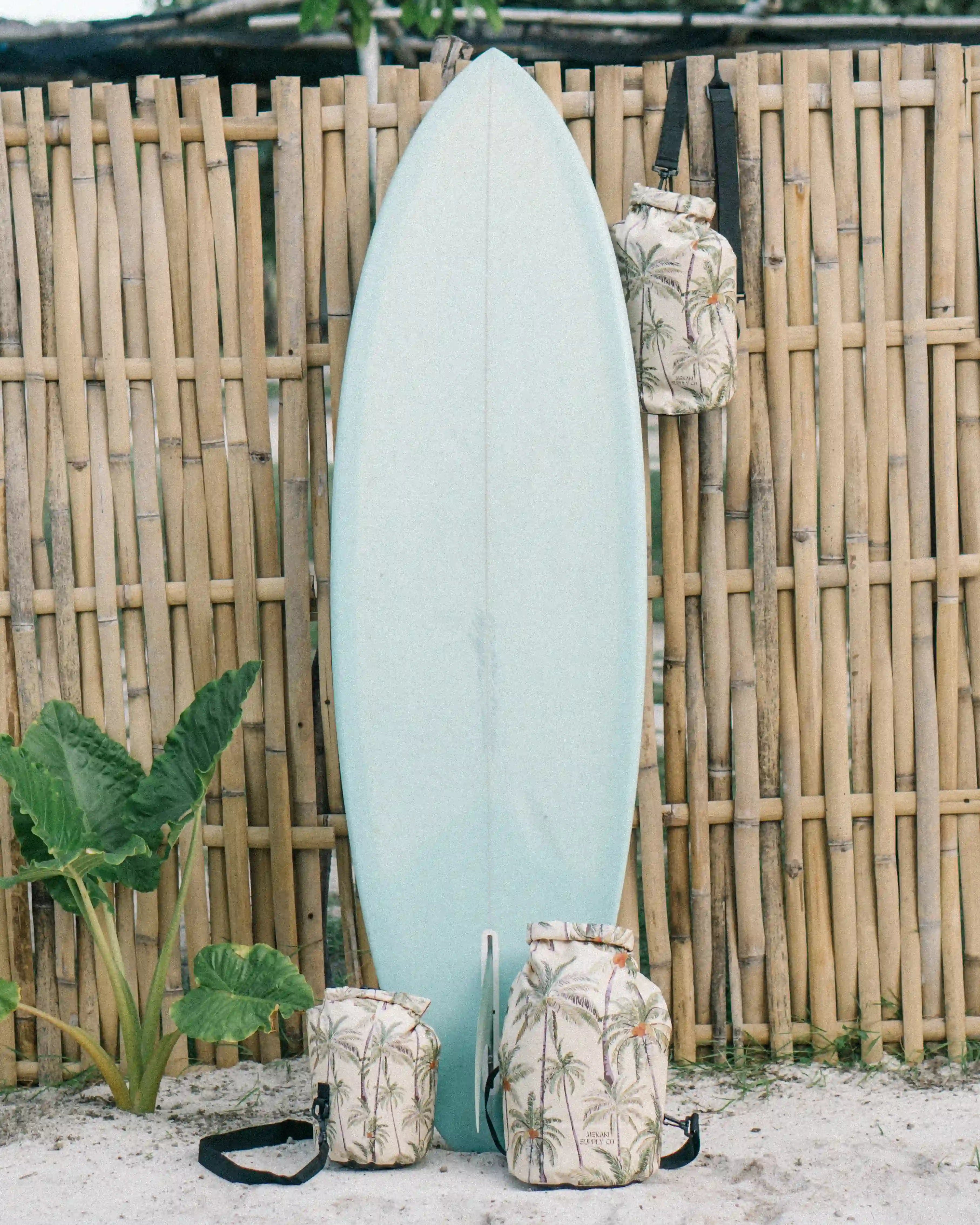 Ein hellblaues Surfbrett lehnt an einem Bambuszaun. Drei Main Design Dry Bag von Meraki Supply Co.-Taschen mit Palmenmotiven hängen am Zaun und liegen im Sand. Im Hintergrund sind üppige grüne Pflanzen und Bäume zu sehen.