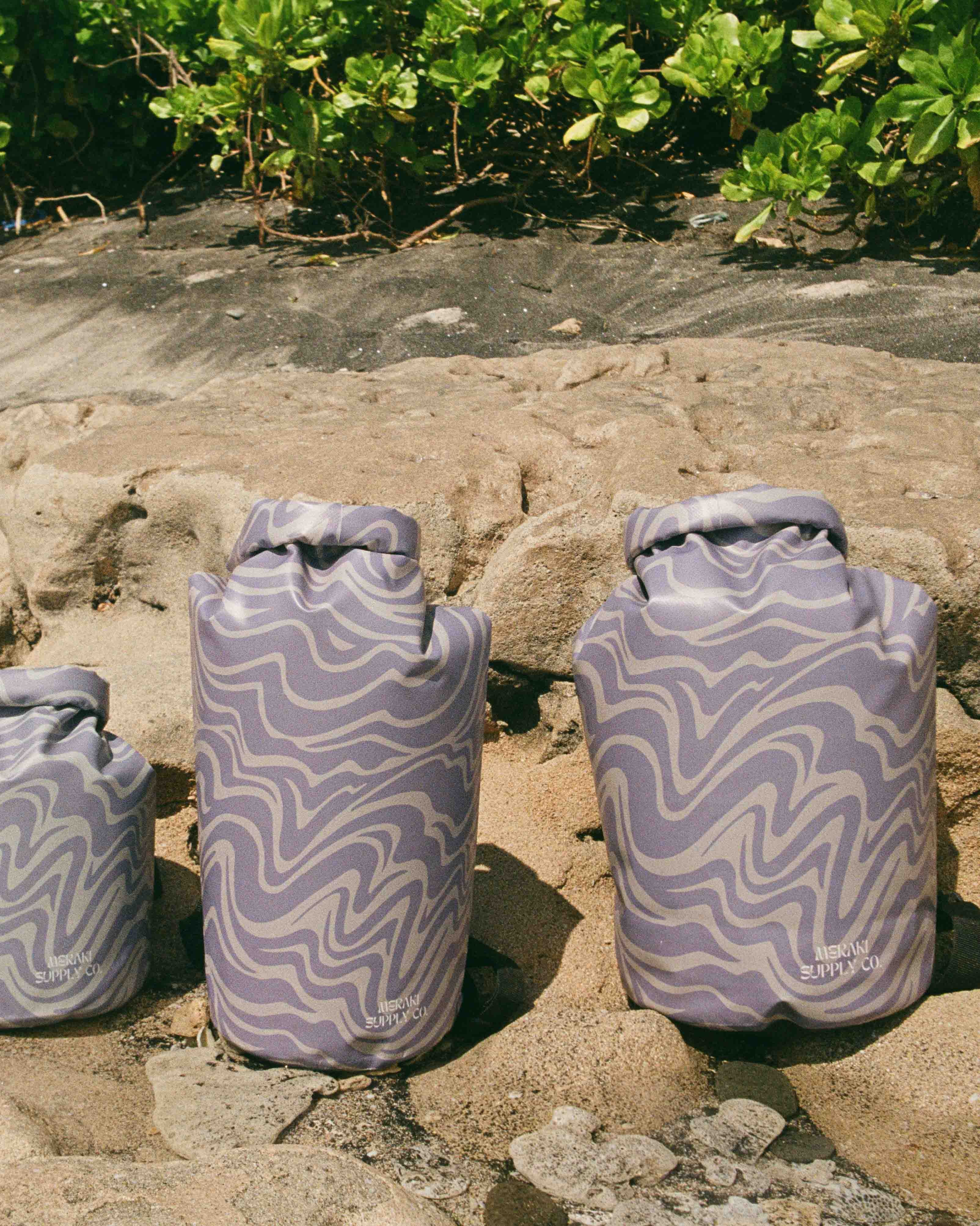 Drei weiche, gemusterte Dry Bag von Meraki Supply Co. by Main Design mit wellenförmigen Designs in Lavendel und Weiß stehen aufrecht auf sandigem Boden – ideal, um Ihre Sachen bei jedem Abenteuer wasserdicht zu halten.