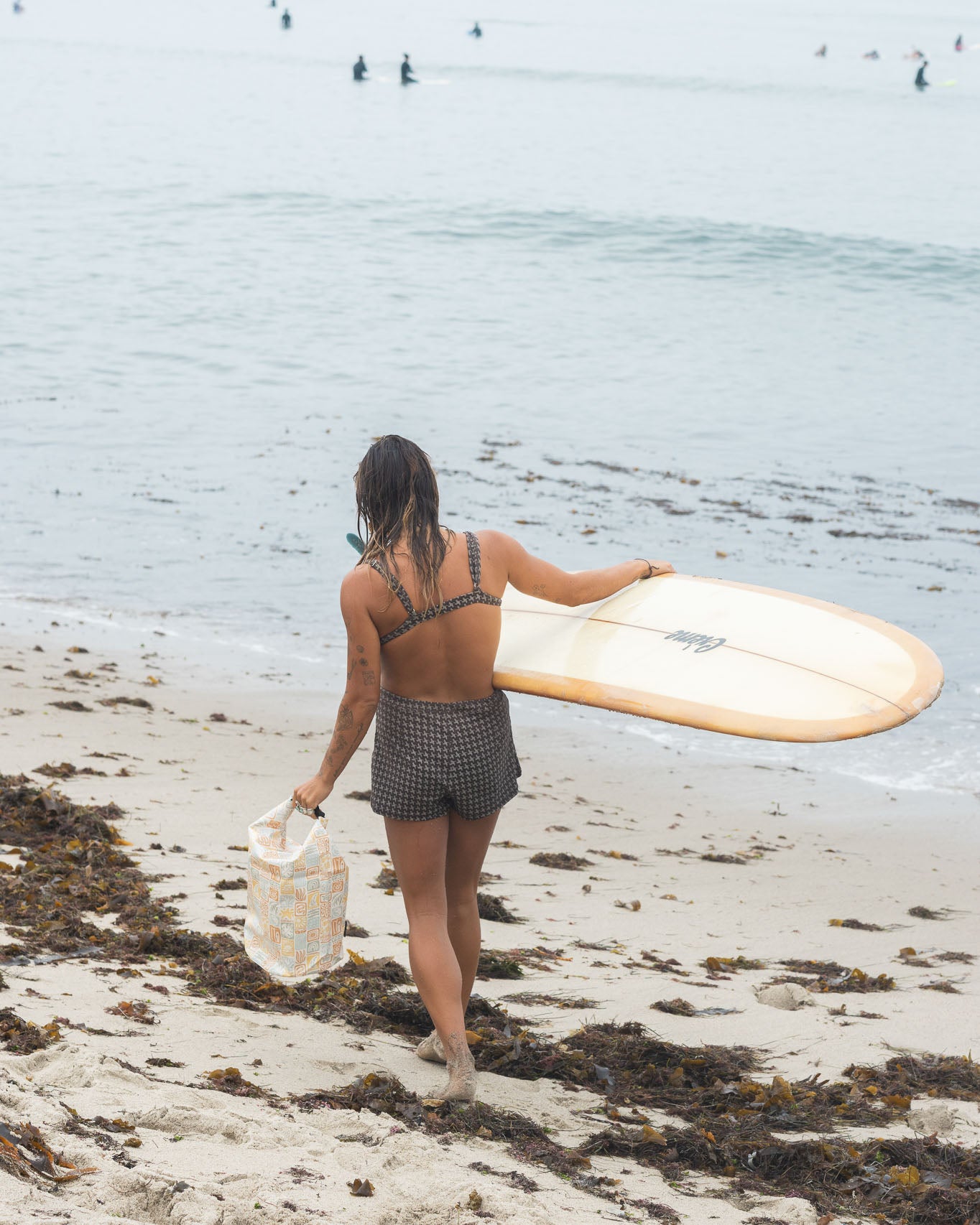 Eine Person in Badebekleidung und Shorts geht über einen mit Algen bedeckten Strand in Richtung Meer. Sie trägt ein Surfbrett und einen Dry Bag von Meraki Supply Co. von Main Design. Auf dem Wasser sind mehrere Surfer zu sehen.
