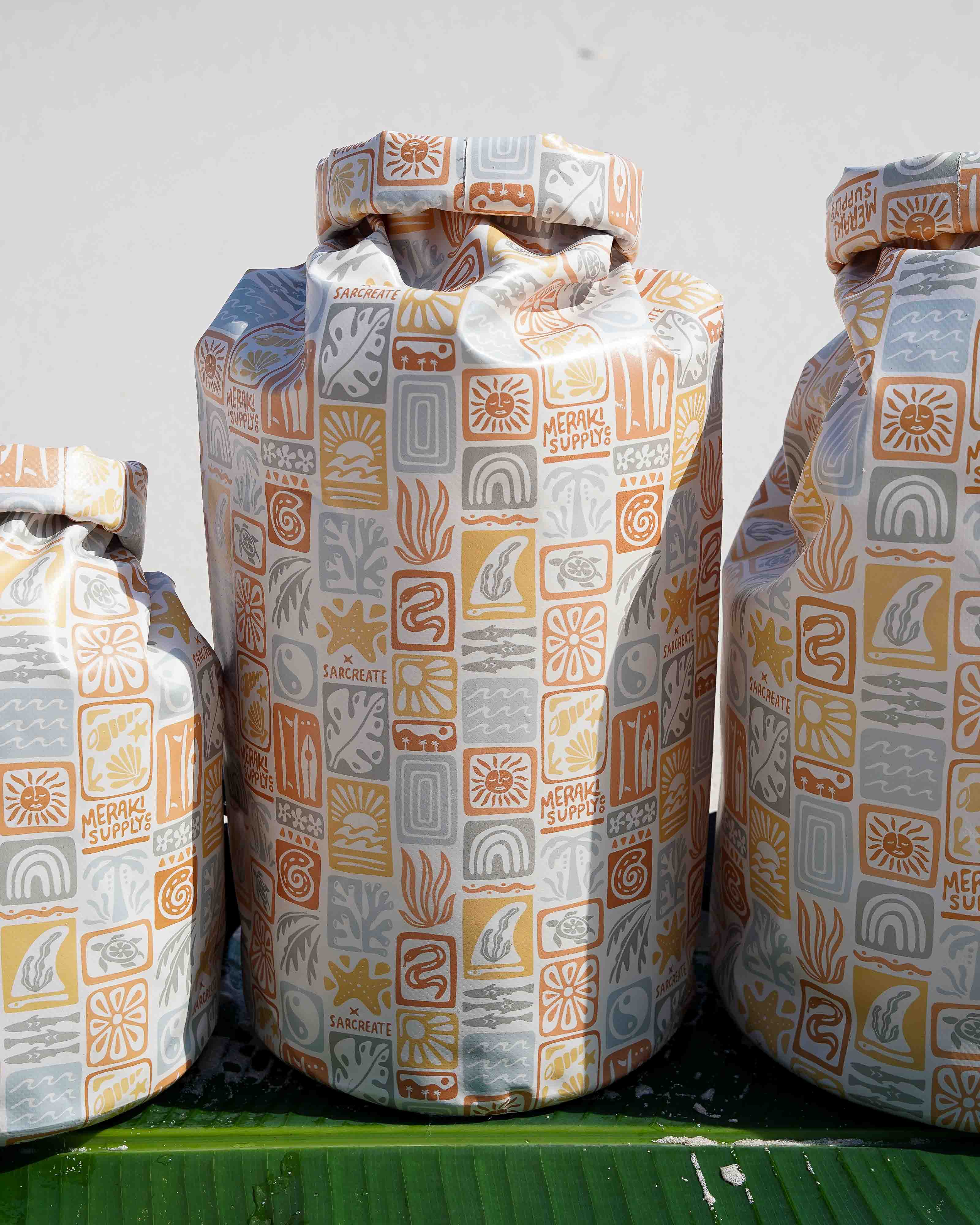 Drei große Dry Bag von Meraki Supply Co. by Main Design mit pastellfarbenen geometrischen und illustrativen Motiven – Wellen, Blumen, „SAGRATE“ – stehen aufrecht auf einer grünen Fläche. Die Taschen sind wasserdicht und stilvoll gemustert.