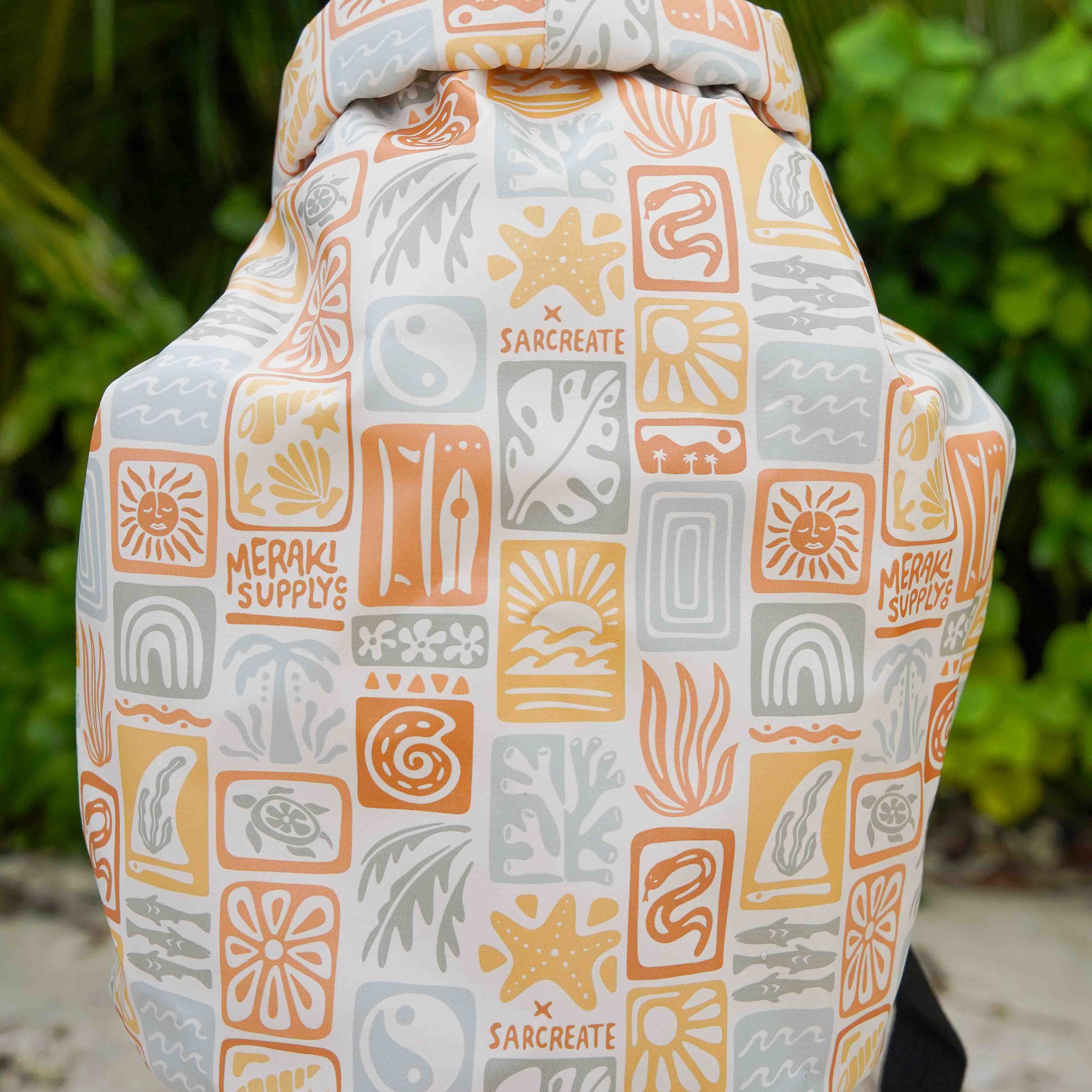 Eine Nahaufnahme eines farbenfrohen, wasserdichten Dry Bag von Meraki Supply Co. von Main Design mit von der Natur inspirierten Mustern und Markenzeichen vor üppigen grünen tropischen Pflanzen.