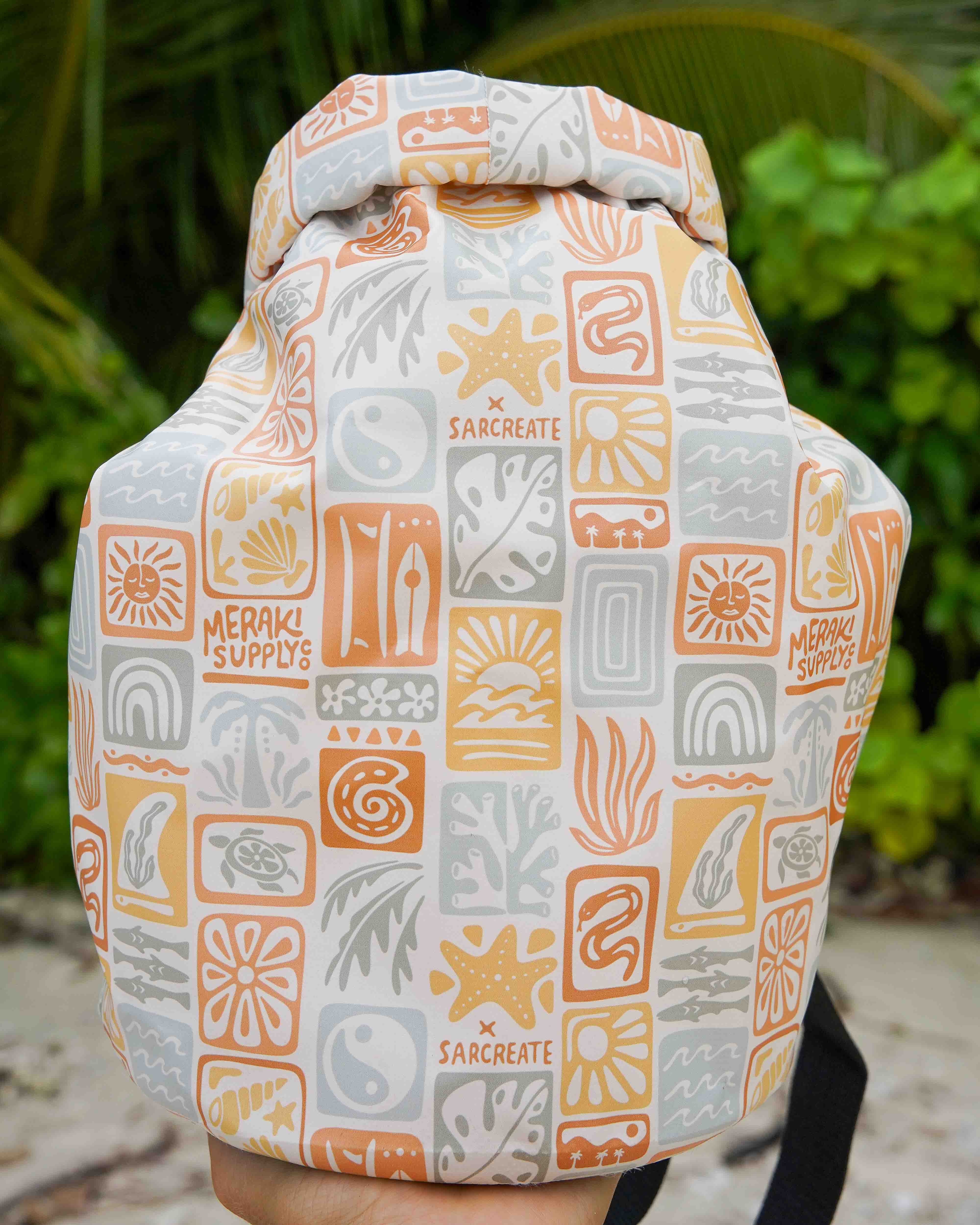 Eine Nahaufnahme eines farbenfrohen, wasserdichten Dry Bag von Meraki Supply Co. von Main Design mit von der Natur inspirierten Mustern und Markenzeichen vor üppigen grünen tropischen Pflanzen.