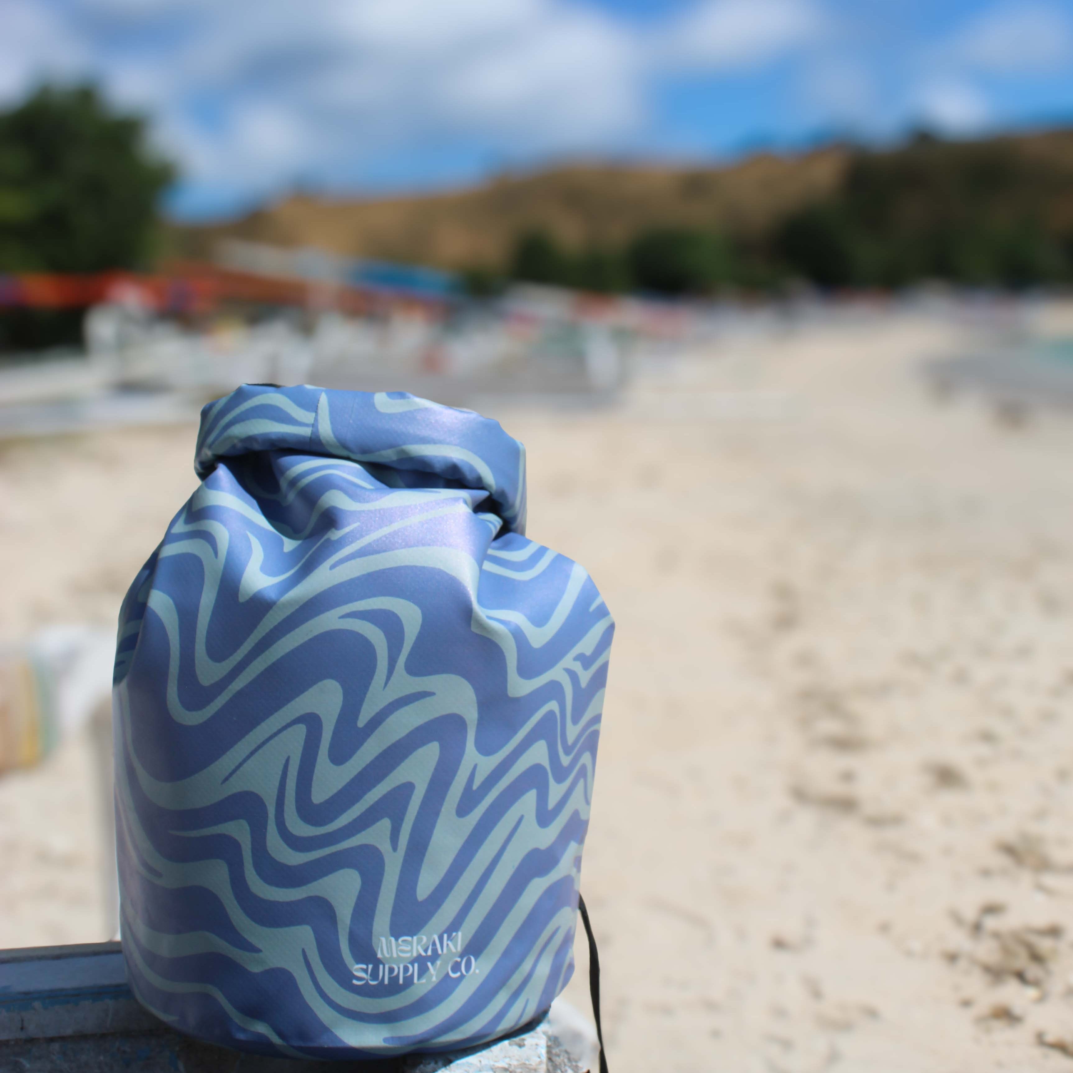 Ein Dry Bag von Meraki Supply Co. by Main Design mit Wellenmuster und wasserdichtem Material liegt an einem Sandstrand in der Nähe eines blauen Seils mit verschwommenen Bäumen, Booten und Hügeln im teilweise bewölkten Hintergrund.