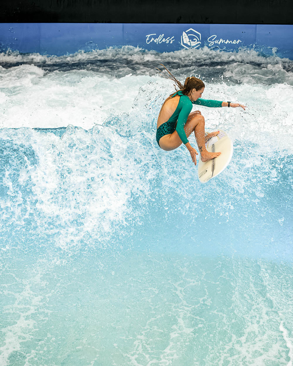 Ein Surfer im Main Design Eclipse Surf Suit reitet auf einer Welle im Haus, die Knie gebeugt und in der Luft über dem glitzernden blauen Wasser, während weiße Gischt aufsteigt und "Endless Summer" an die Wand dahinter geschrieben steht.