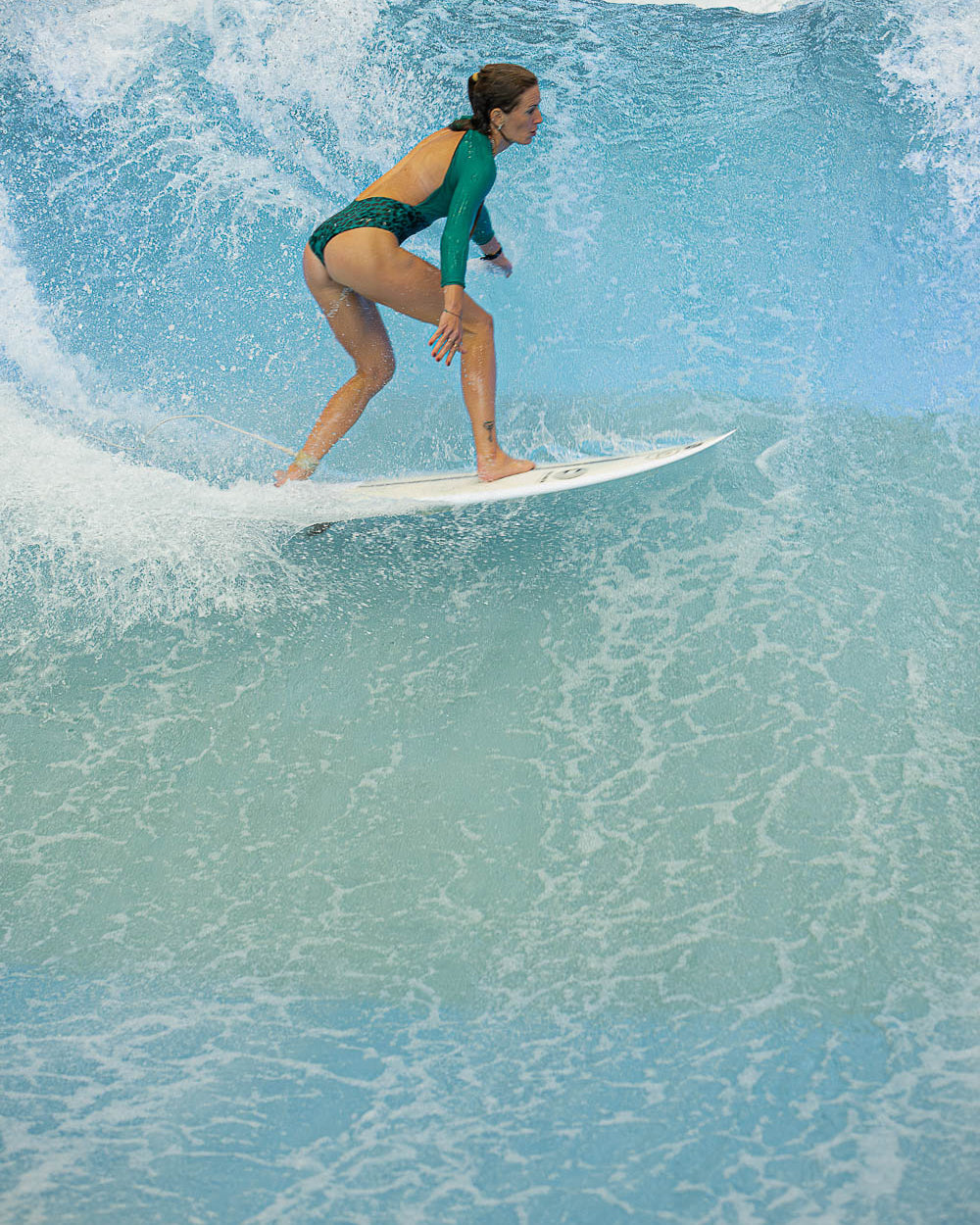 Eine Frau, die den Main Design Eclipse Surf Suit aus nachhaltigem Material trägt, surft auf einer großen blauen Welle und balanciert auf einem weißen Surfbrett, während das Wasser um sie herum plätschert.