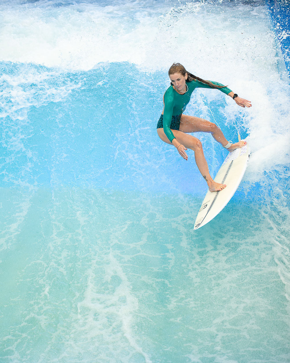 Eine Frau in einem grünen Main Design Eclipse Surf Suit reitet auf einem weißen Surfbrett auf einer Welle und lehnt sich in die Kurve, während das Wasser um sie herum in einem hellblauen Pool spritzt.