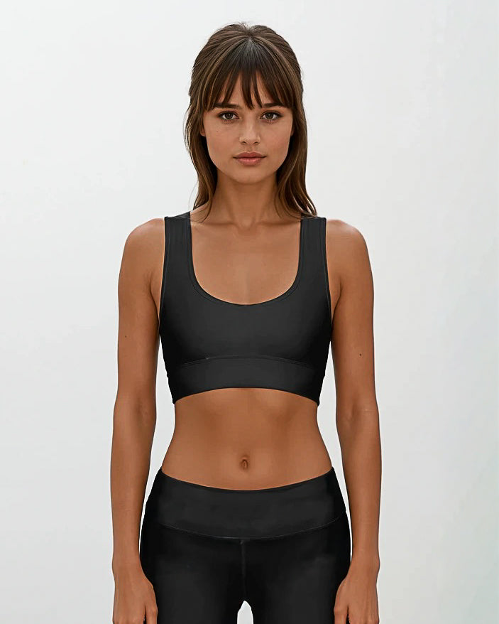 Eine Frau mit langen braunen Haaren und Pony trägt das Main Design Flow Active Top und schwarze Leggings. Sie steht vor einem schlichten hellen Hintergrund und schaut mit neutralem Blick direkt in die Kamera.