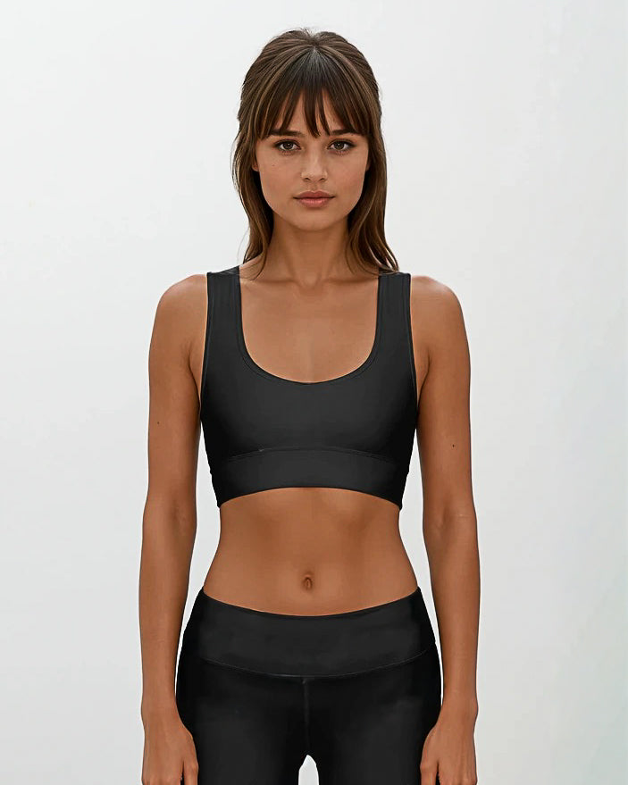 Eine Frau mit langen braunen Haaren und Pony trägt das Main Design Flow Active Top und schwarze Leggings. Sie steht vor einem schlichten hellen Hintergrund und schaut mit neutralem Blick direkt in die Kamera.