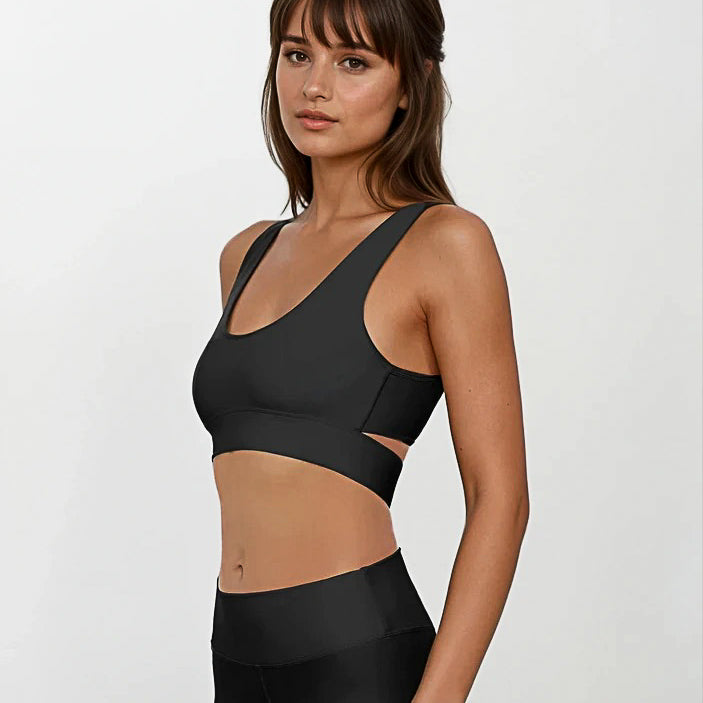 Eine Frau mit glattem braunem Haar und Pony trägt das Main Design Flow Active Top und schwarze Leggings mit hoher Taille aus recycelten Materialien, steht vor einem schlichten hellen Hintergrund und blickt mit neutralem Blick in die Kamera.