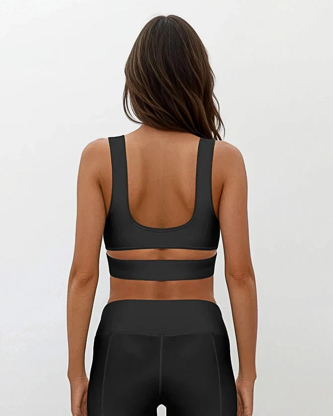 Eine Frau mit langen braunen Haaren blickt in die Ferne. Sie trägt das Main Design Flow Active Top mit einem breiten horizontalen Träger und dazu passende Leggings mit hoher Taille vor einem schlichten hellen Hintergrund.