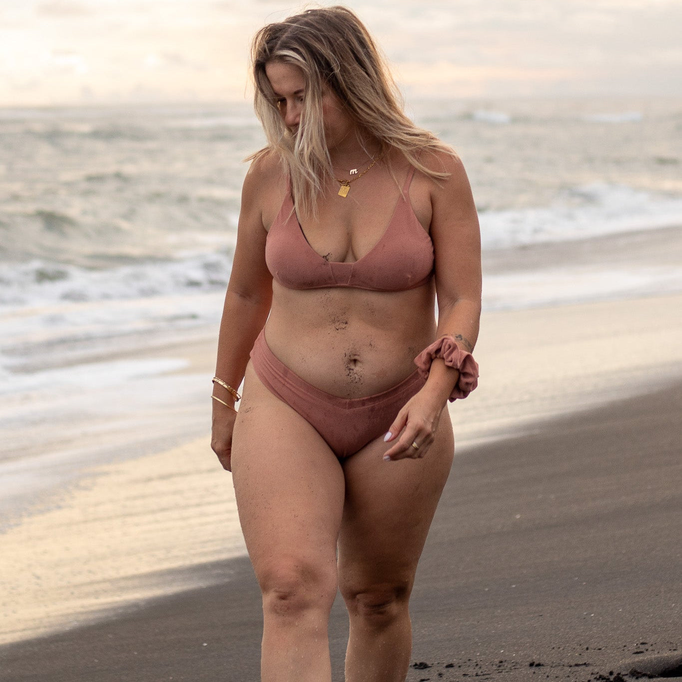 Eine Frau mit langen Haaren spaziert in der wendbaren Gem-Bikinihose von Main Design am Strand entlang. Die Meereswellen brechen sanft unter einem bewölkten Himmel, während sie mit einem Haargummi am Handgelenk nach unten blickt und über ihre Wahl nachhaltiger Bademode nachdenkt.