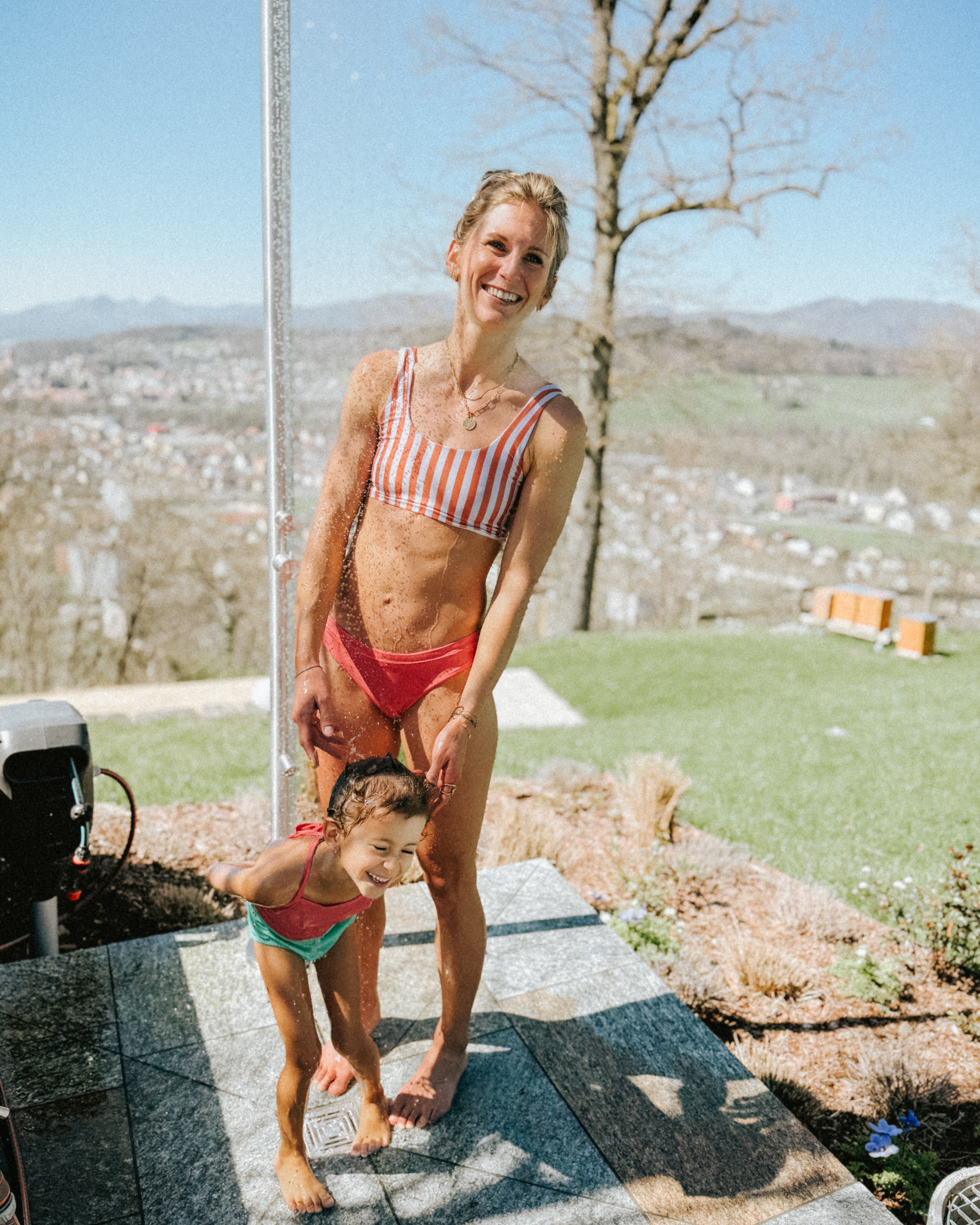 Eine lächelnde Frau trägt das Hale Bikini Oberteil von Main Design mit Rundhalsausschnitt und steht mit einem Kind in Badebekleidung draußen unter der Dusche. Sie genießen die Sonne, eingerahmt von Bäumen und einer malerischen Stadt in der Ferne im Hintergrund.