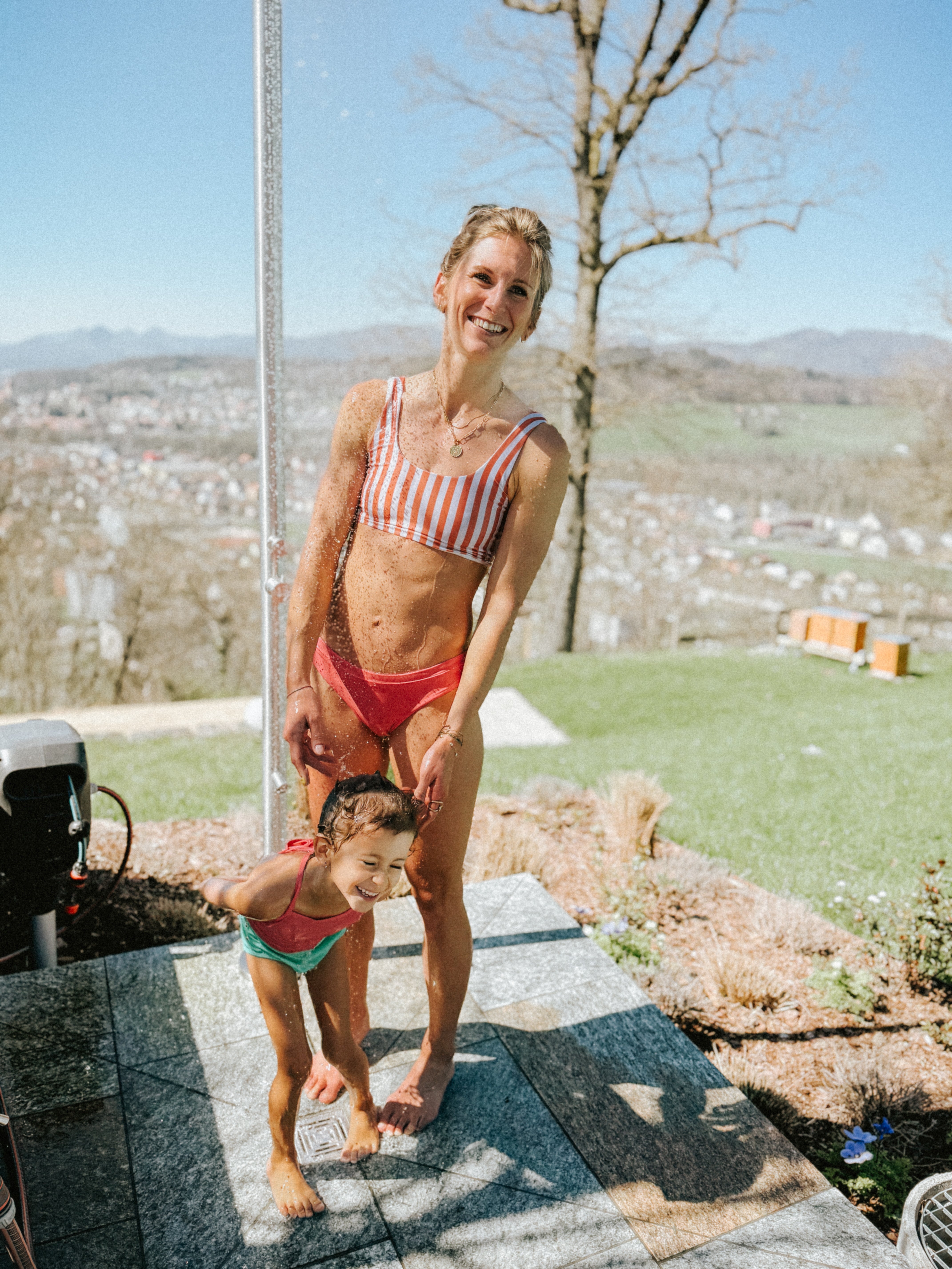 Eine lächelnde Frau trägt das Hale Bikini Oberteil von Main Design mit Rundhalsausschnitt und steht mit einem Kind in Badebekleidung draußen unter der Dusche. Sie genießen die Sonne, eingerahmt von Bäumen und einer malerischen Stadt in der Ferne im Hintergrund.