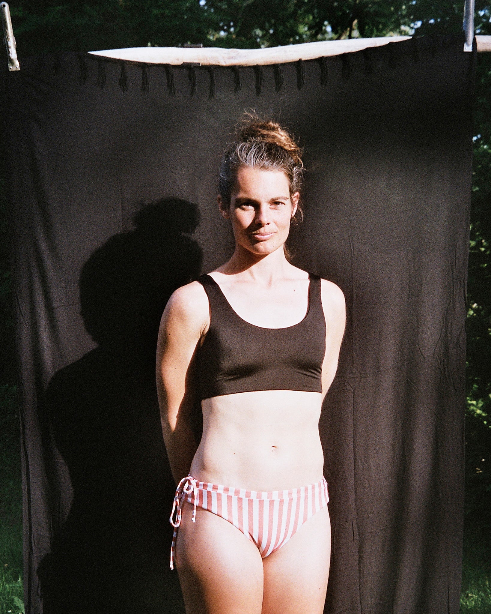 Eine Frau steht im Freien vor einem schwarzen Stoffhintergrund und trägt das Main Design Hale Bikini-Oberteil in Schwarz mit rundem Ausschnitt und rosa-weiß gestreiftem Bikiniunterteil. Im Hintergrund sind Bäume und Grünpflanzen zu sehen.