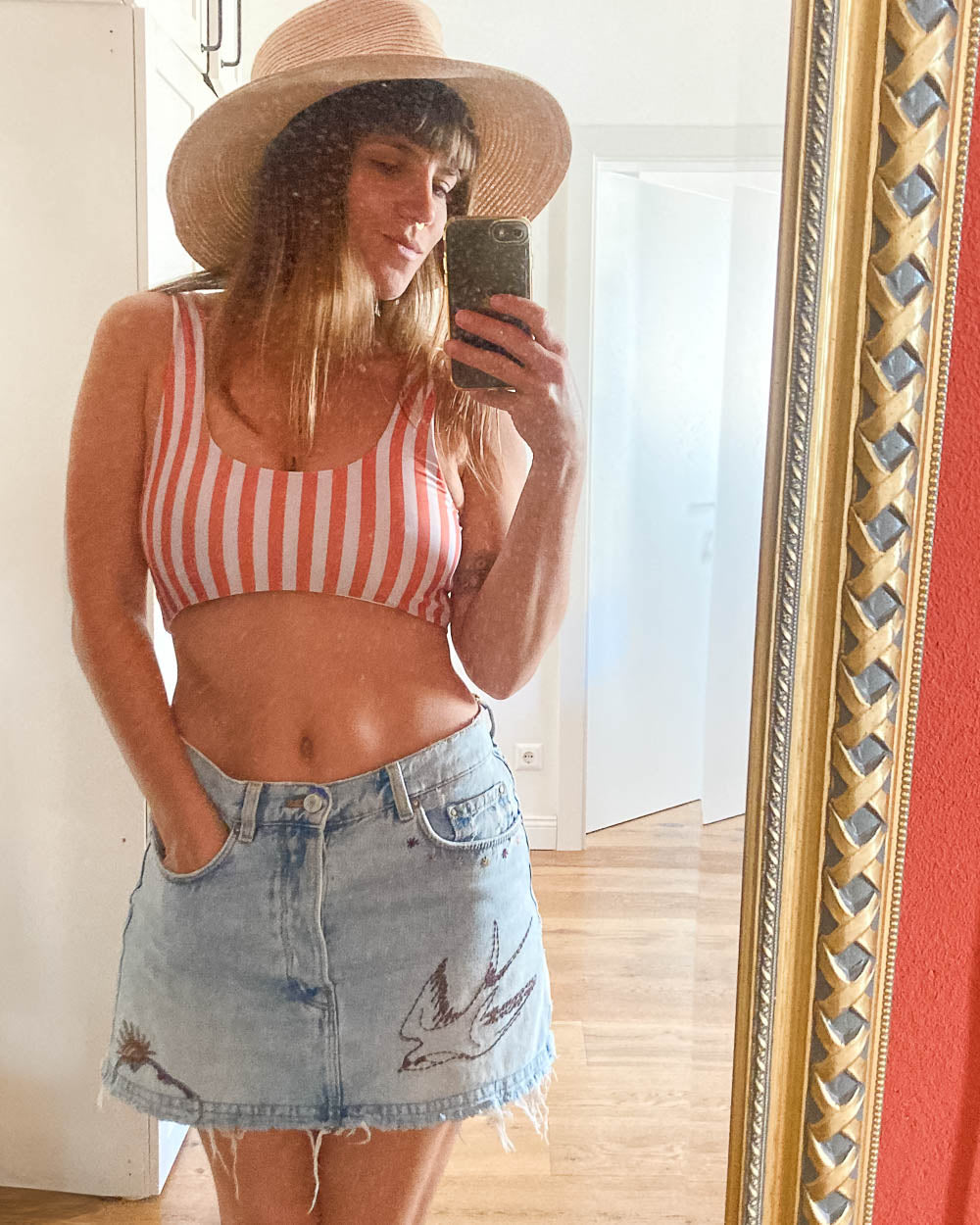 Eine Frau in einem hellen Raum mit Holzfußboden macht ein Selfie im Spiegel. Sie trägt das Main Design Hale Bikini Oberteil und einen leichten Jeansrock mit Vogelmotiven, dazu einen breitkrempigen Hut.
