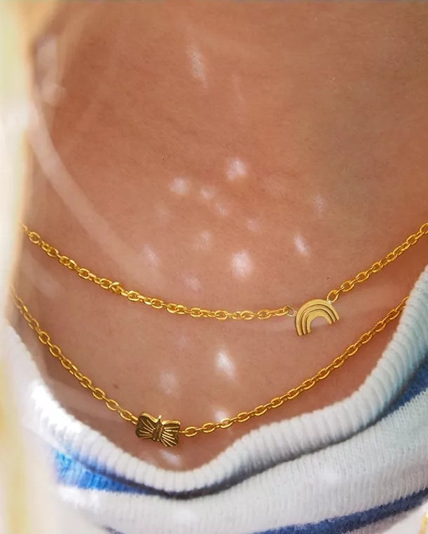 Nahaufnahme einer Person, die zwei zarte Adorabili Halsketten aus 24 Karat vergoldetem Gold - eine mit einem Schmetterlingsanhänger, eine mit einem Regenbogenanhänger - über einem blau-weiß gestreiften Hemd trägt. Das Sonnenlicht schimmert auf der Haut. Ideal als Geschenk für Kinder.