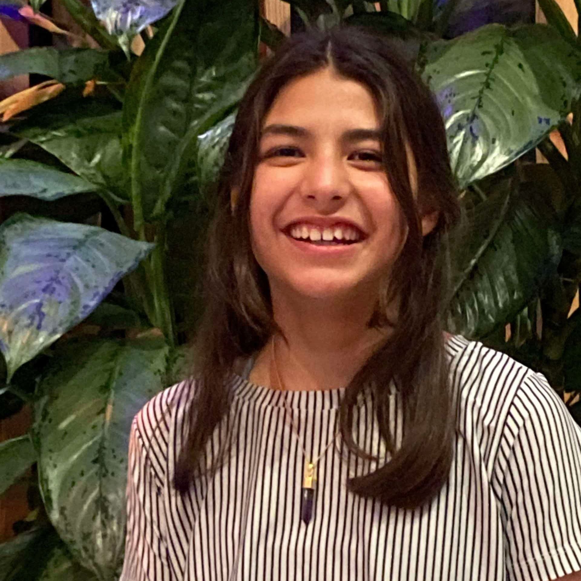 Eine junge Person mit langen dunklen Haaren, die ein gestreiftes Hemd und eine Halskette trägt, lächelt vor großen grünen Blättern in einem Innenraum.