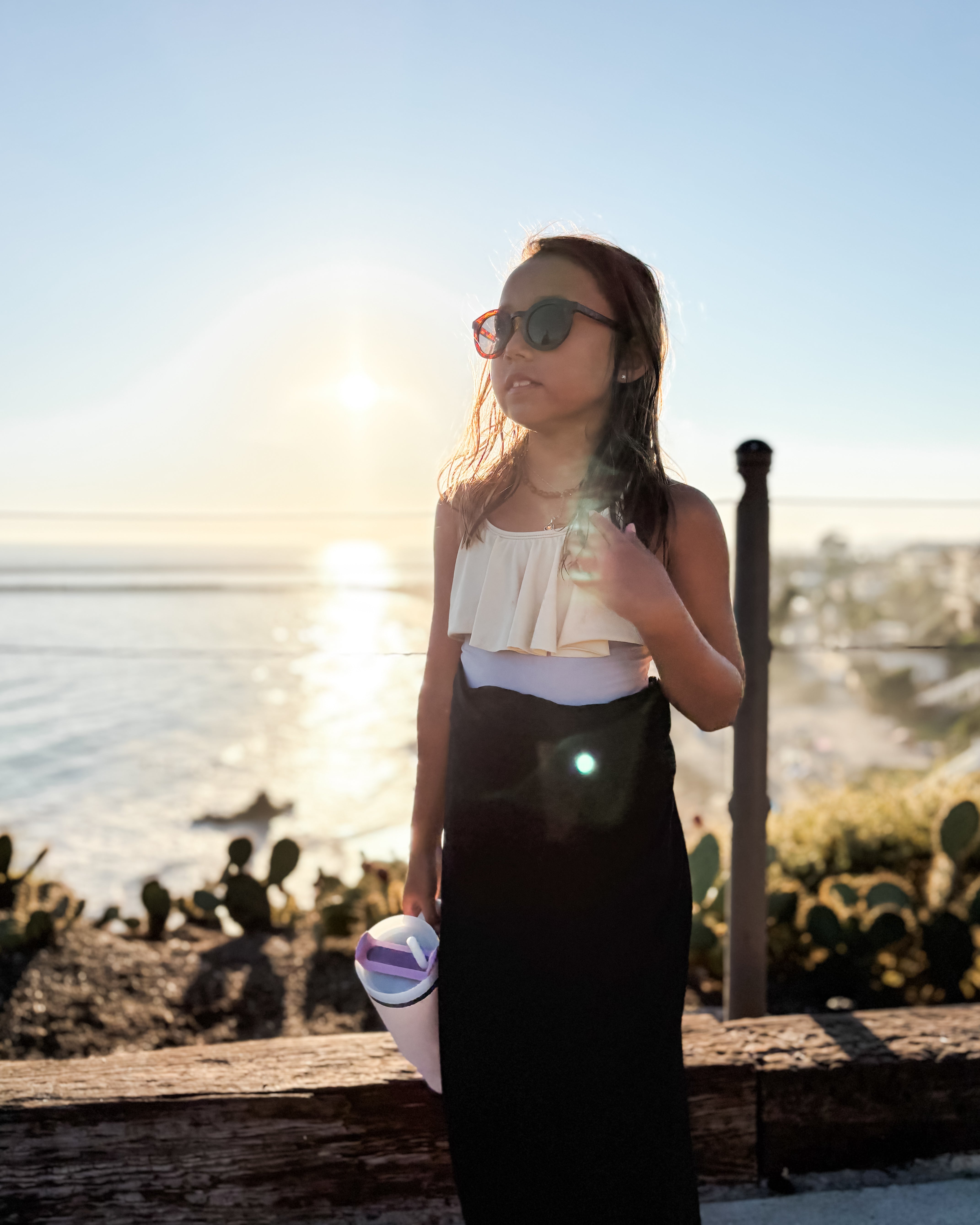 Ein junges Mädchen mit Sonnenbrille und einem Sarong Bali von Main Design steht bei Sonnenuntergang am Meer und hält eine Tasse in der Hand, während die Sonne hinter ihr leuchtet, sich auf dem Wasser spiegelt und ein warmes Licht auf die Szene wirft.