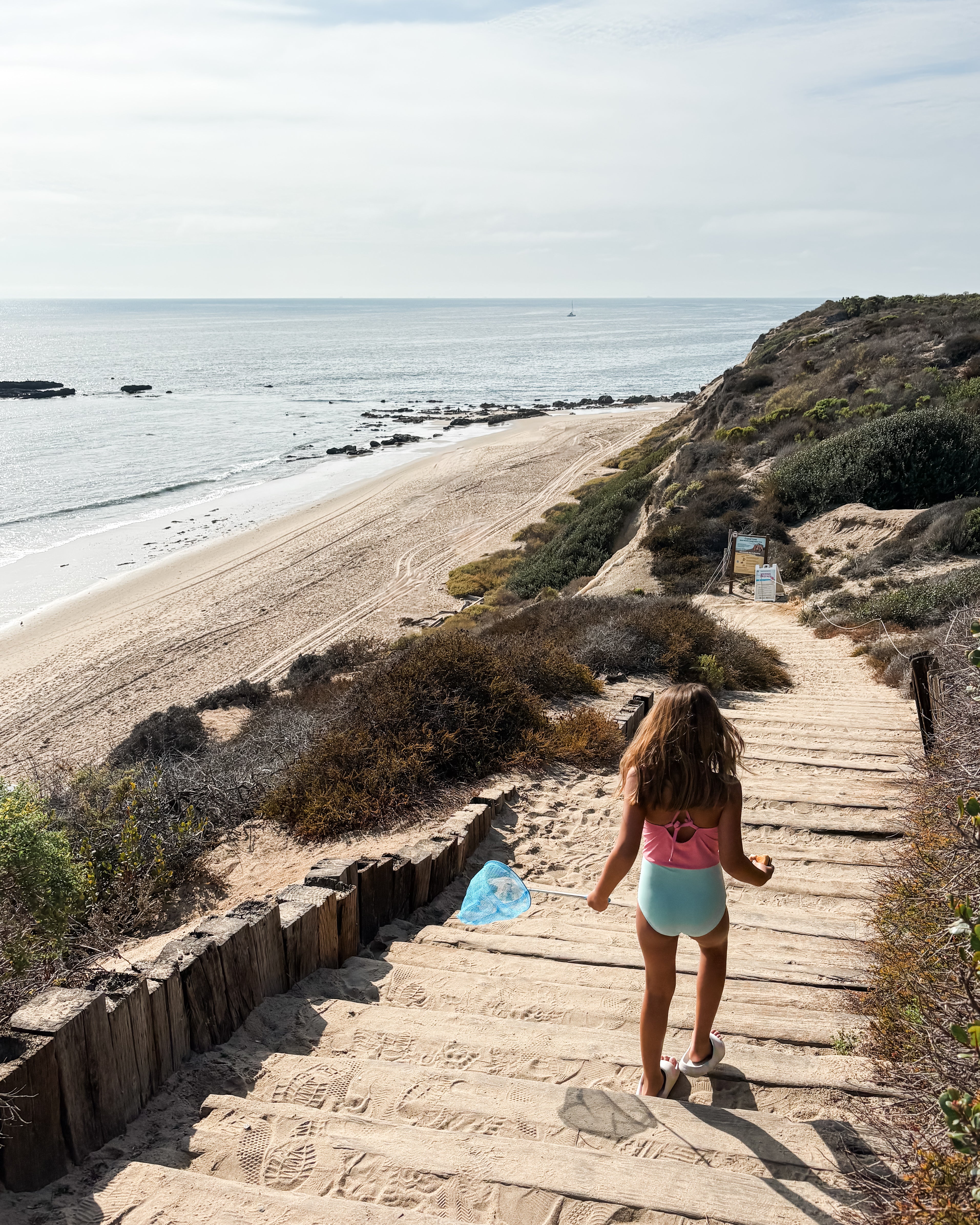 Ein Mädchen im Lena Mädchen Badeanzug von Main Design aus recycelten Materialien läuft über sandige Holzstufen zum ruhigen Strand, mit Meer, Felsenküste und bewachsenen Klippen unter klarem Himmel.