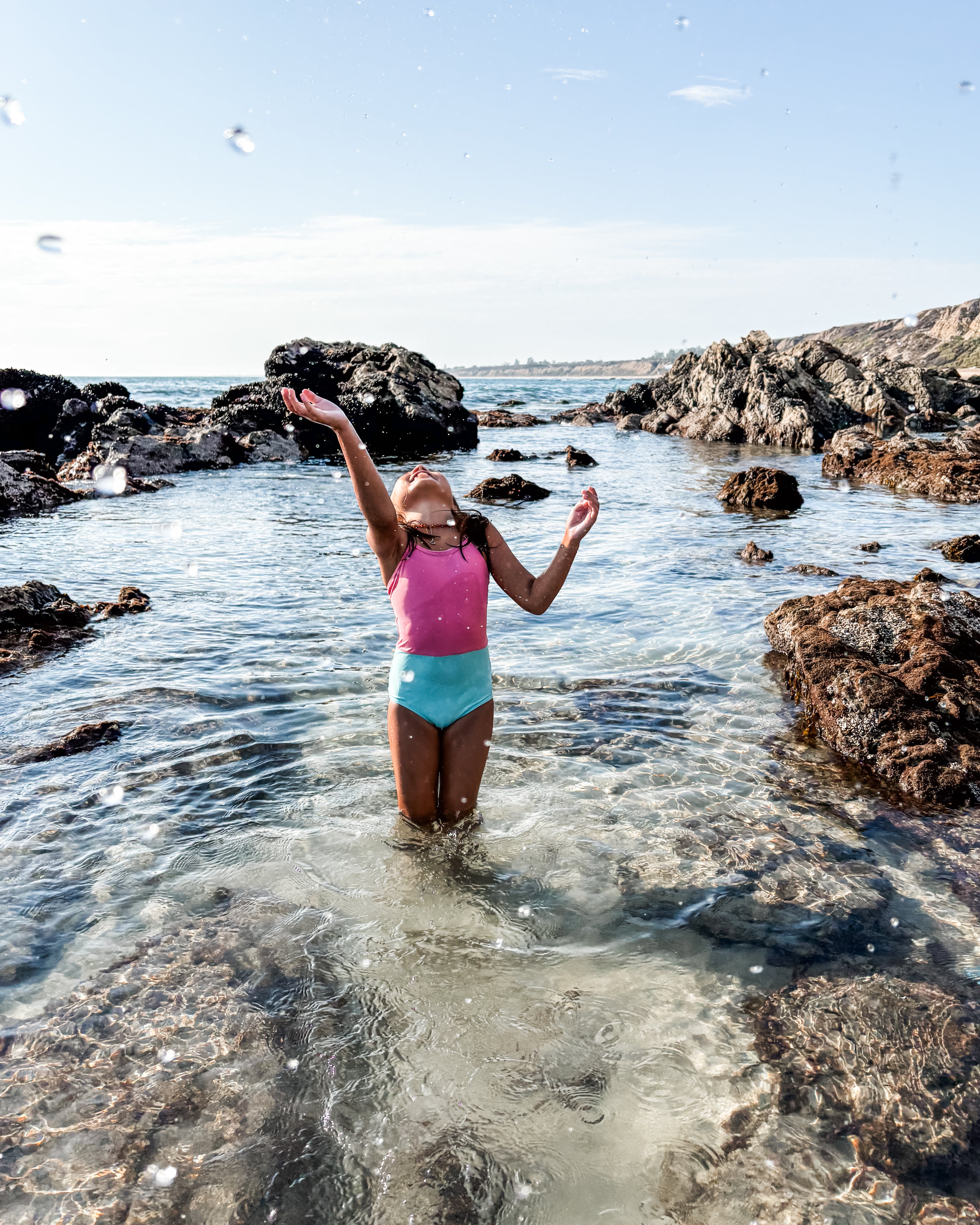 Ein Kind in einem Lena Mädchen Badeanzug von Main Design, rosa und türkis, steht knietief im klaren, felsigen Meerwasser und streckt sich freudig nach oben, mit Felsen und einer entfernten Küstenlinie im Hintergrund.