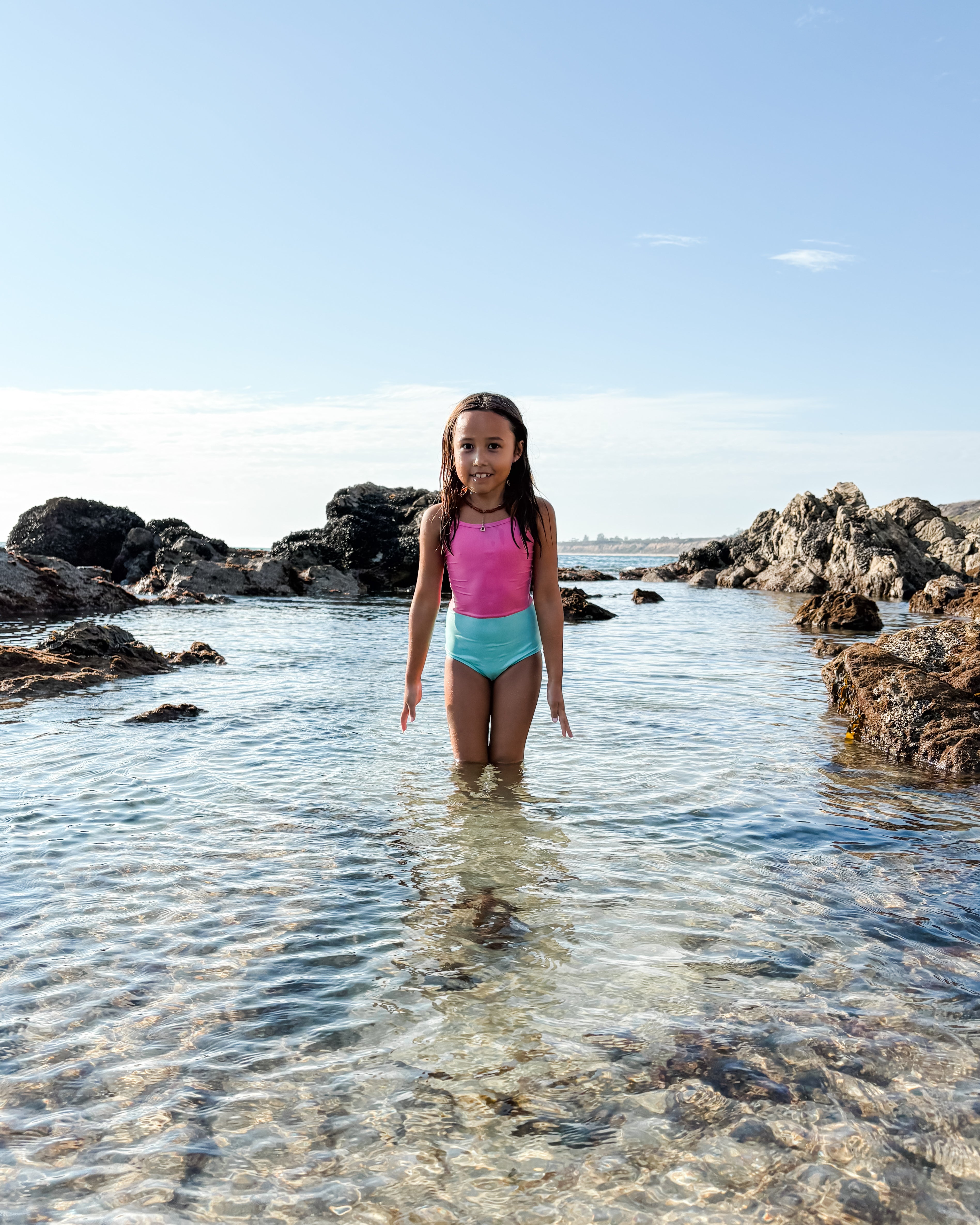 Ein junges Mädchen steht im klaren, flachen Wasser zwischen Felsen am Strand und trägt den Lena Mädchen Badeanzug von Main Design aus recycelten Materialien in Pink und Blau. Ein heller Himmel und eine ferne Küstenlinie vervollständigen die Szene.