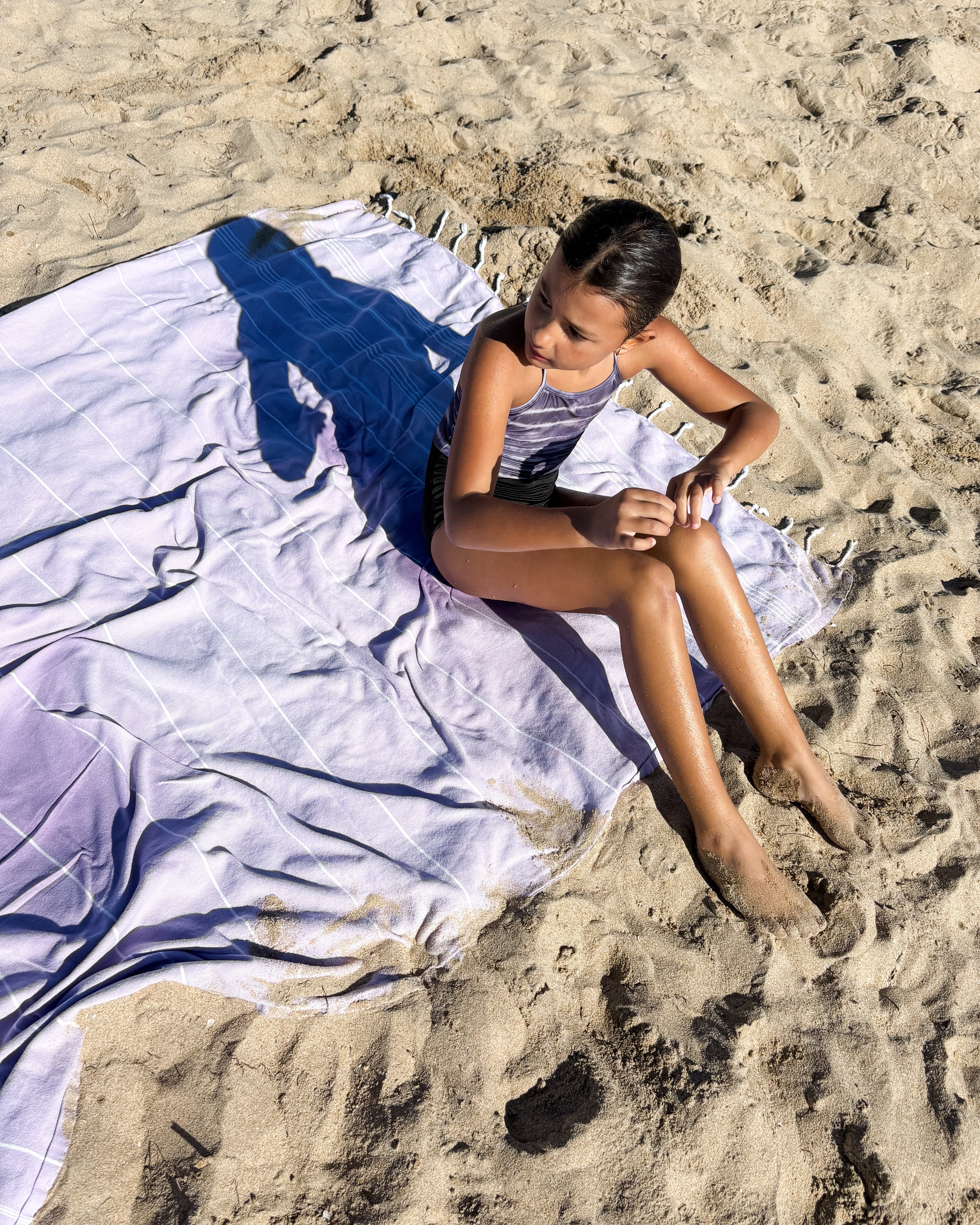 Ein junges Mädchen im Badeanzug sitzt auf einem großen, hellvioletten Maxi Strandtuch De La Mer von Main Design, das am Sandstrand ausgebreitet ist. Das Sonnenlicht wirft ihren Schatten auf das Handtuch, während sie mit ausgestreckten sandigen Füßen zur Seite schaut.