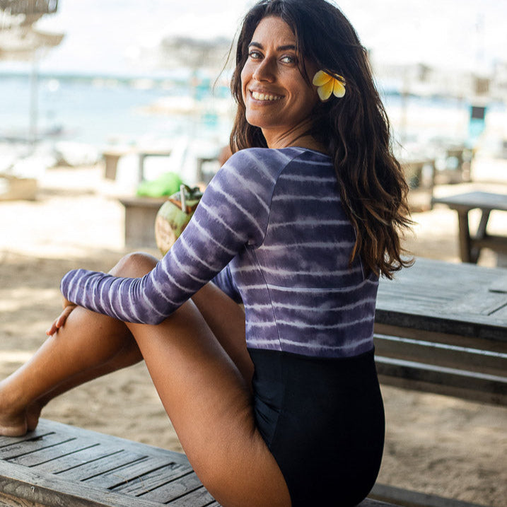 Eine Frau sitzt seitlich auf einer Holzbank am Strand und lächelt in die Kamera. Sie hat eine Blume im Haar und trägt einen Ivy Surf Suit von Main Design und schwarze Shorts aus recycelten Materialien. Der Strand ist mit Lichterketten dekoriert, im Hintergrund stehen Tische und Sonnenschirme.