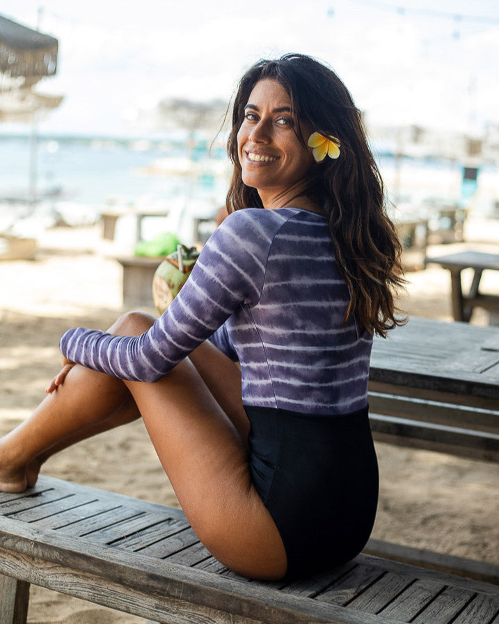 Eine Frau sitzt seitlich auf einer Holzbank am Strand und lächelt in die Kamera. Sie hat eine Blume im Haar und trägt einen Ivy Surf Suit von Main Design und schwarze Shorts aus recycelten Materialien. Der Strand ist mit Lichterketten dekoriert, im Hintergrund stehen Tische und Sonnenschirme.