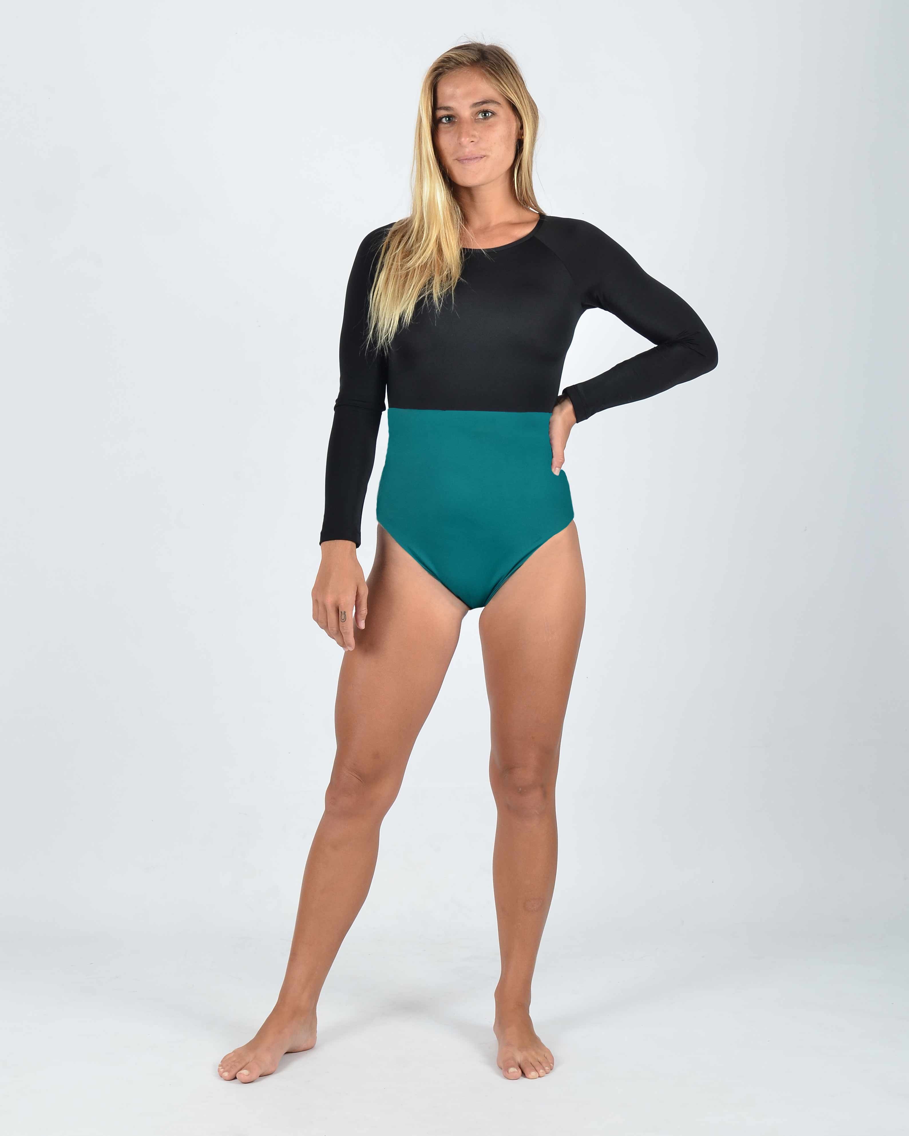 Eine Frau mit langen blonden Haaren steht barfuß vor einem schlichten Hintergrund. Sie trägt den Main Design Ivy Surf Suit in Schwarz und Blaugrün aus recycelten Materialien. Ihre linke Hand ruht auf der Hüfte, ihr rechter Arm hängt herab. Dieser nachhaltige Anzug bietet LSF 50+.