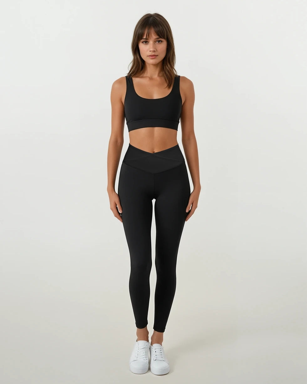 Eine Frau blickt vor einem schlichten weißen Hintergrund nach vorne. Sie trägt die Juno Leggings von Main Design aus recyceltem Nylon mit hoher Taille, kombiniert mit einem schwarzen Sport-BH und weißen Turnschuhen.