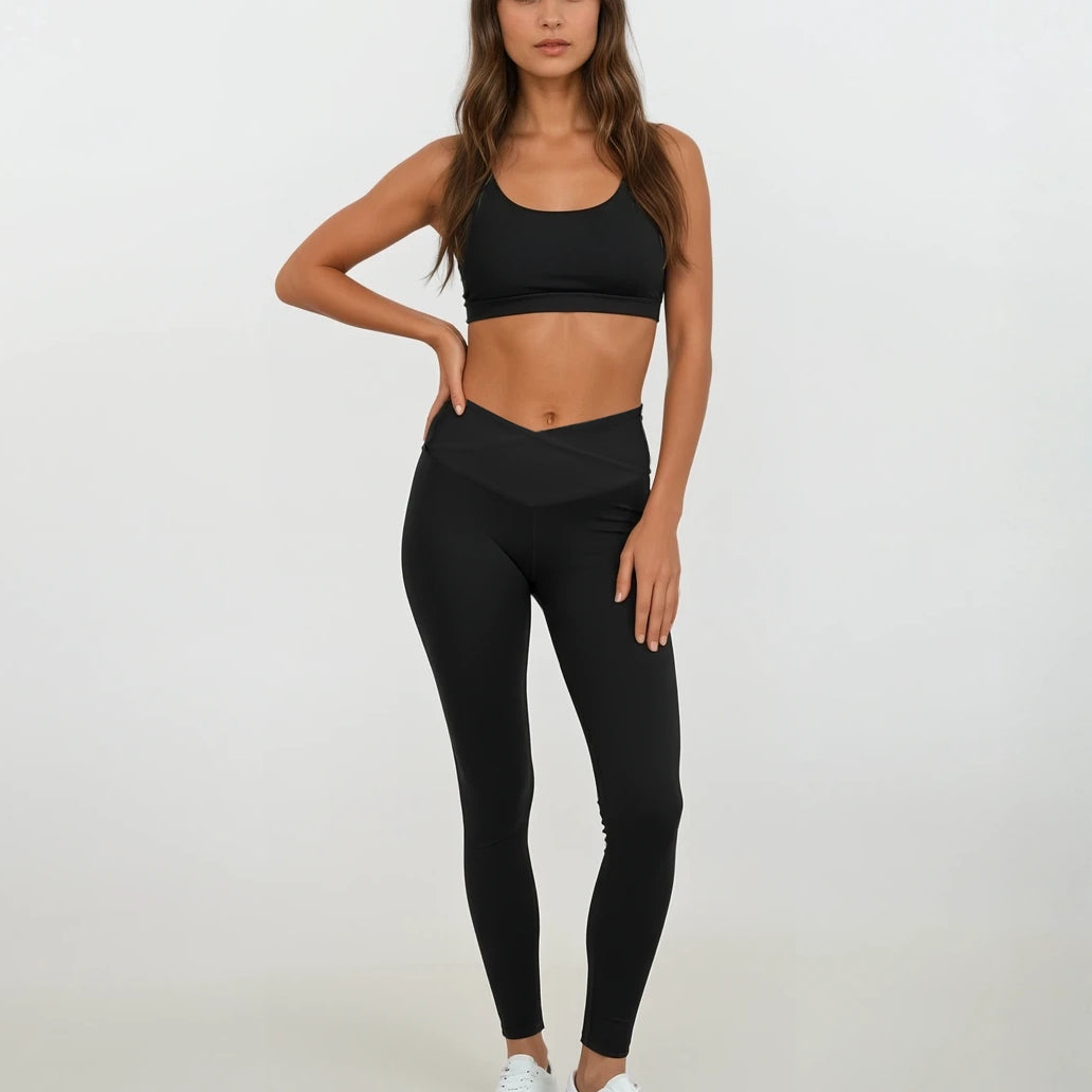 Eine Frau mit langen braunen Haaren trägt Main Design Juno Leggings mit hohem Bund aus recyceltem Nylon, einen schwarzen Sport-BH und weiße Sneakers vor weißem Hintergrund. Ihre linke Hand liegt auf der Hüfte.