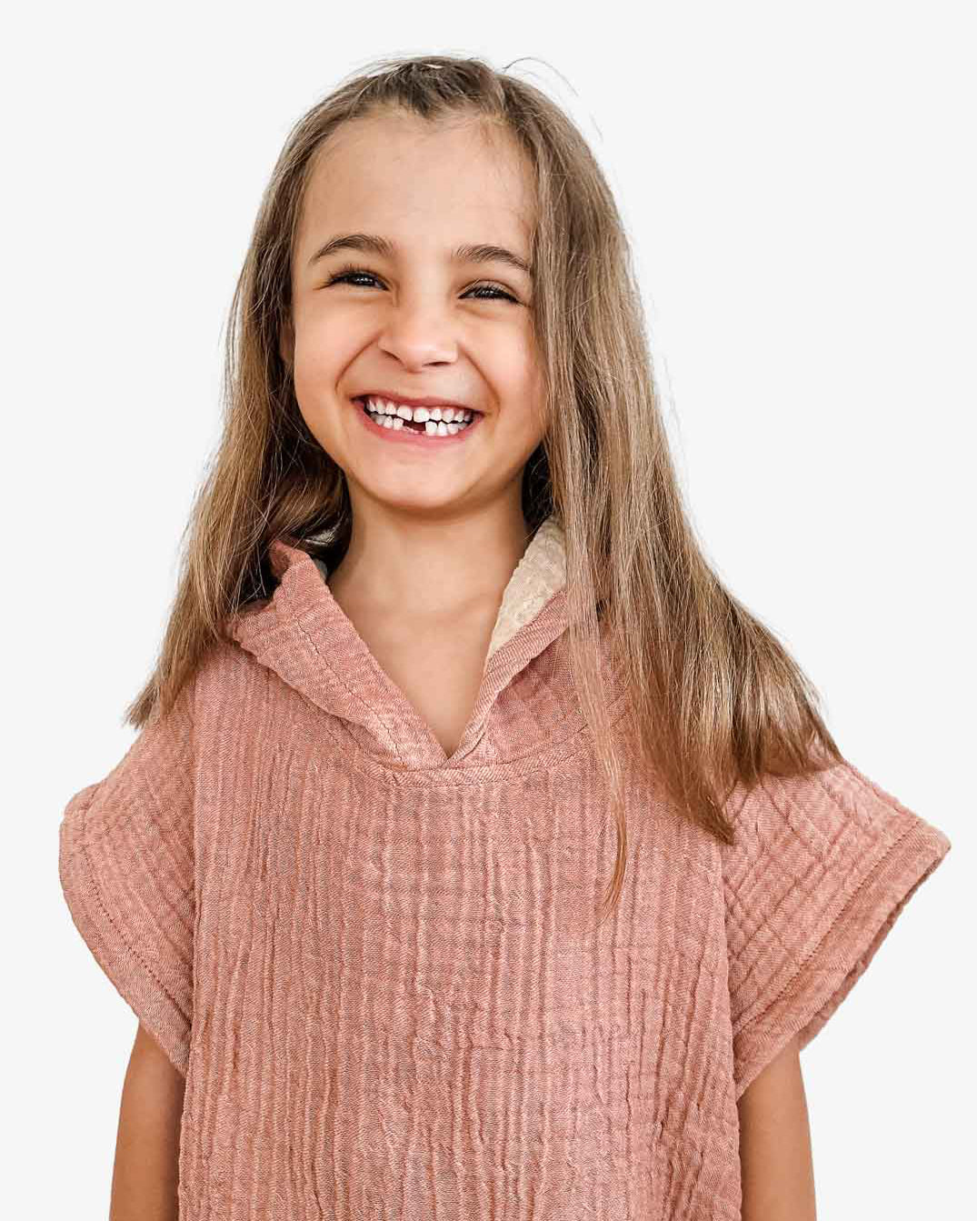 Ein lächelndes Kind mit langen braunen Haaren trägt den „Kinder Badeponcho Flow“ von Main Design aus OEKO-TEX zertifizierter Baumwolle. Der strukturierte rosa Musselinstoff verleiht dem reinweißen Hintergrund sanfte Eleganz.