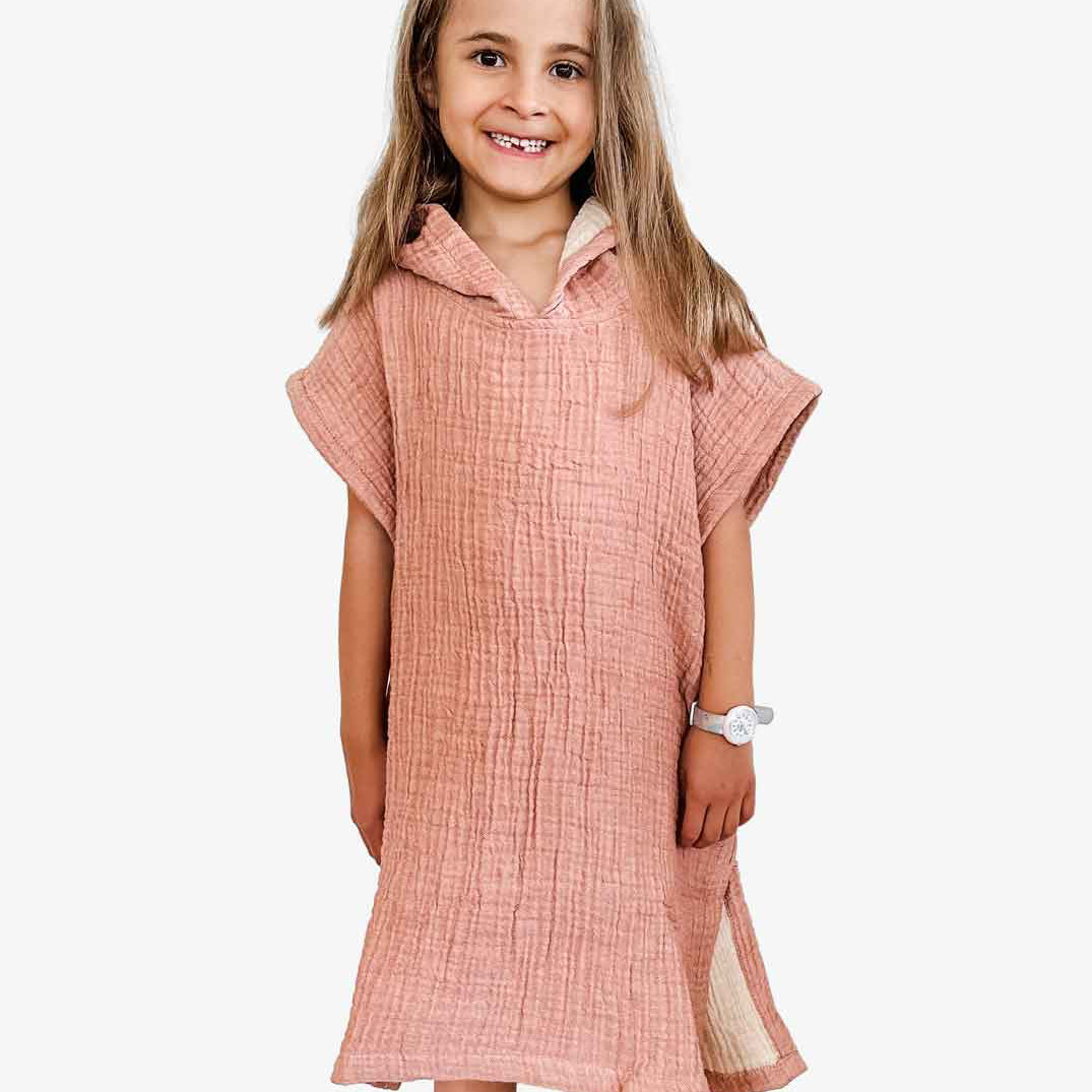 Ein junges Mädchen mit langen Haaren lächelt und trägt einen Main Design Kinder Badeponcho Flow in Pink. Sie ist aus OEKO-TEX zertifizierter Baumwolle gefertigt, steht barfuß vor einem schlichten weißen Hintergrund und trägt eine Uhr am linken Handgelenk.