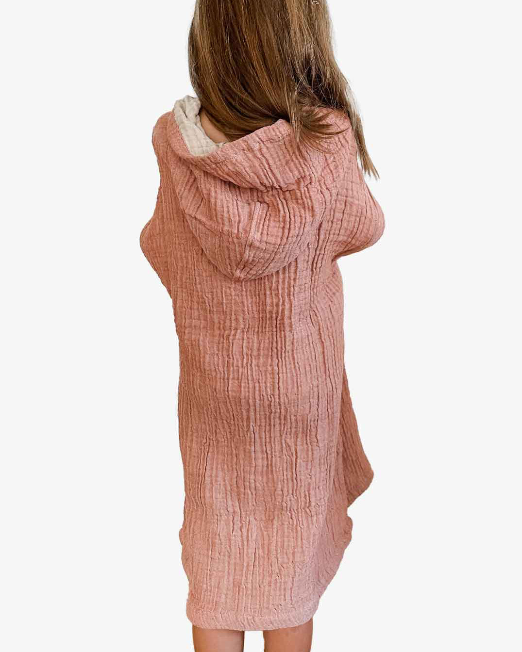 Ein Kind mit langen Haaren, von hinten gesehen, ist kuschelig in einen rosa Main Design Kinder Badeponcho Flow aus OEKO-TEX zertifizierter Baumwolle gehüllt. Der strukturierte Stoff fällt bis zum Knie vor einem schlichten weißen Hintergrund.