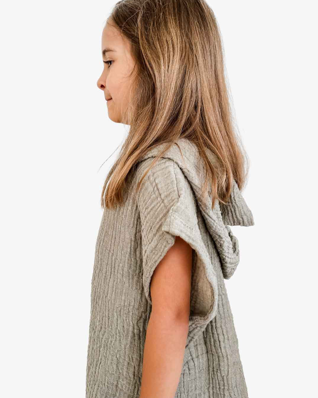 Ein junges Mädchen mit langen braunen Haaren steht im Profil, eingehüllt in einen Main Design Kinder Badeponcho Flow aus OEKO-TEX zertifizierter Baumwolle. Die hellgrüne Kapuze liegt über ihren Schultern, während der schlichte weiße Hintergrund die Textur des Musselinstoffs betont.